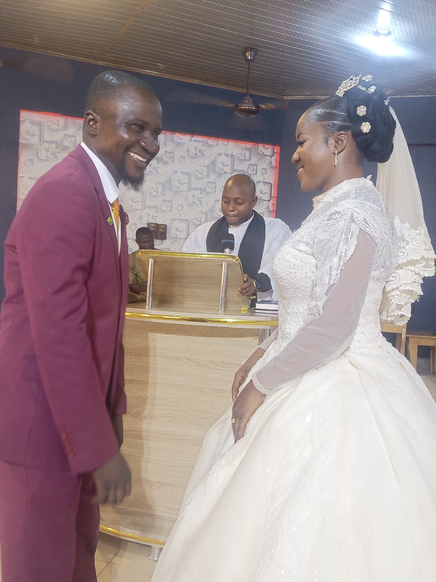 Here's a detailed description of the image:
**Scene:** A wedding ceremony likely taking place in a church or religious building in Akwanga, Nigeria.
**People:**
* A groom wearing a burgundy suit with a yellow tie is seen on the left, smiling broadly and looking towards the bride.
* A bride is on the right, dressed in a traditional white wedding gown with long, sheer sleeves and a full skirt. She is also smiling and looking at the groom. Her hair is styled in an updo with floral ornaments and a veil.
* An officiant is standing behind a wooden lectern with gold trim, likely conducting the ceremony. He is wearing what appears to be a religious vestment.
* A second person is visible in the background on the left, behind the lectern.
**Objects:**
* Wooden lectern with gold trim.
* Microphone
* Building interior with painted walls, ceiling fan, and fluorescent lights.
**Actions/Interactions:**
* The groom and bride are exchanging glances and smiling, suggesting a moment of joy and connection during the wedding ceremony.
* The officiant is engaged in the ceremony.
**Environment:**
* The scene is indoors, presumably within a church or religious building. The background is somewhat blurred, but it appears to be a simple interior. The lighting is artificial and overhead.