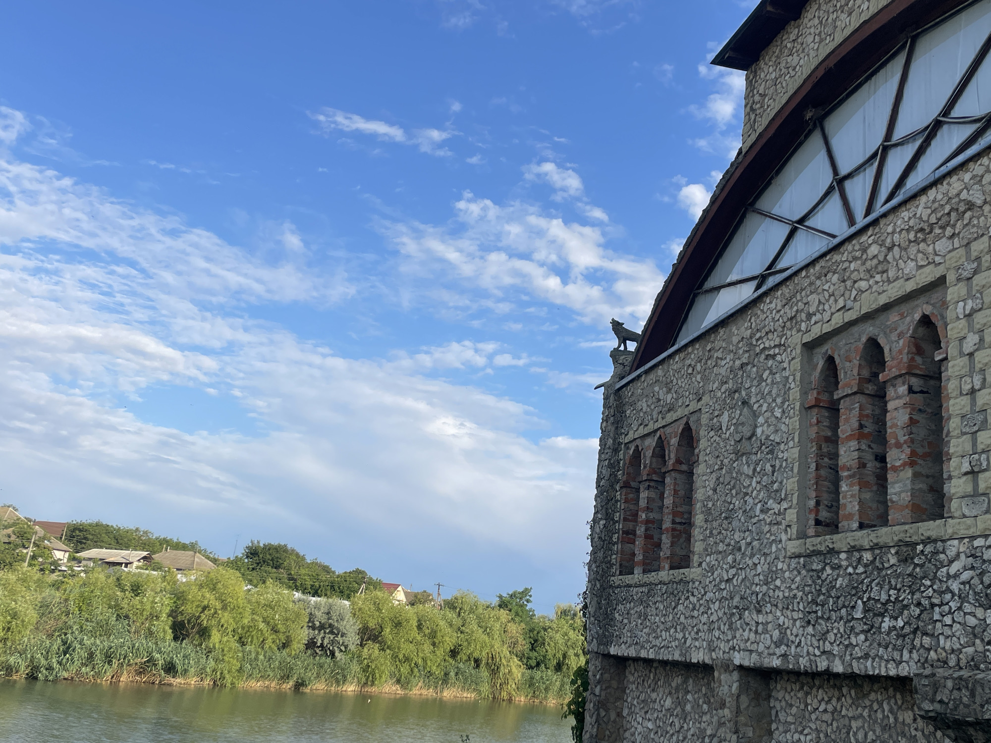 A stone building with arched windows and a large triangular window is situated next to a body of water. A small statue of a dog stands on the roof of the building. The water is bordered by lush green vegetation, and in the background, there are houses and trees. The sky is blue with scattered white clouds. The image is captured from a slightly elevated perspective, looking across the water towards the building and the opposite shore.