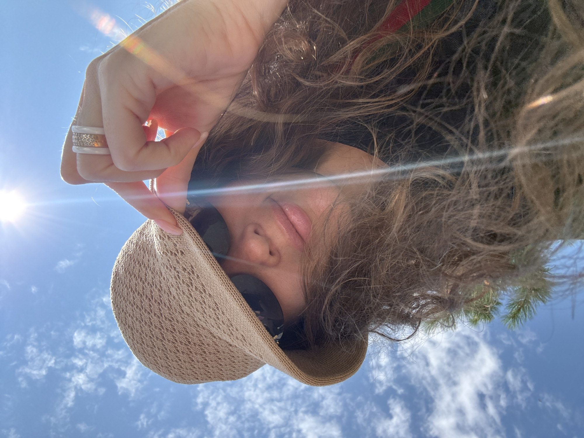 In Artybash, Russia, under a bright blue sky with scattered clouds and the sun shining brightly, a woman is captured in a low-angle selfie. She wears a light brown, crocheted bucket hat and dark sunglasses. Her long, wavy brown hair is visible around her face and shoulders, with some strands appearing windblown. She is touching the brim of her hat with her right hand, which is adorned with two rings: one appears to be a white band with gold accents, and the other is a wider white band with a textured gold pattern. Her lips are slightly parted and glossy, and she has a light pink manicure. The lens flare from the sun is prominent, creating a bright streak across the image. In the background, the sky is clear and blue, with wispy white clouds. A hint of green foliage, possibly from a pine tree, is visible on the left side of the frame. The overall impression is a casual, sunlit outdoor moment.