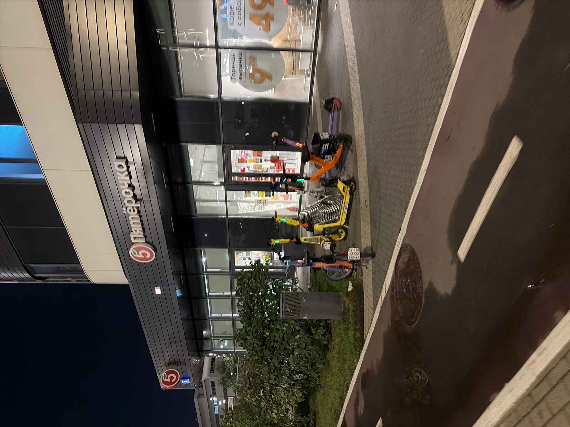 The image depicts the entrance to a Pyaterochka supermarket in Moscow, Russia, at night. The store's facade features a prominent "5 Пятёрочка" logo. In front of the store, a variety of rental electric scooters are parked. Some are yellow, one is orange, and one is purple. A dark grey trash receptacle is situated near a lush green bush. The ground in front of the entrance is paved with dark grey interlocking bricks, and a wet, dark-colored asphalt path with white lane markings runs across the foreground. The overall scene is illuminated by the store's interior lights and ambient external lighting.