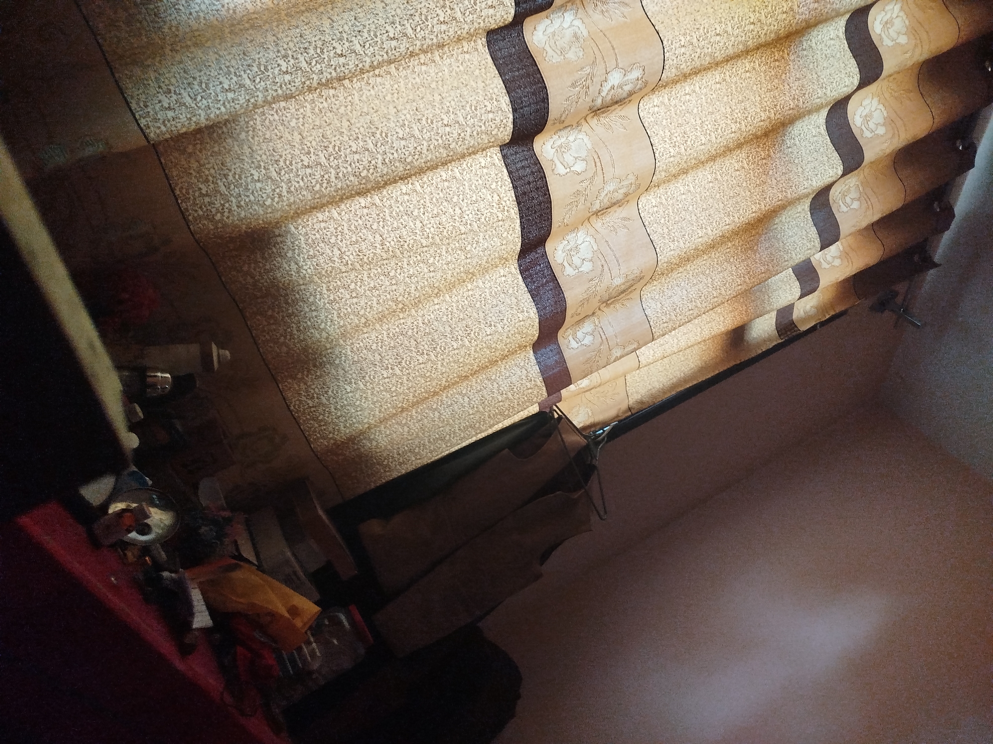 The image is a close-up, tilted shot of a window with horizontal blinds that appear to be made of fabric. The blinds are a light beige color with a textured pattern and a darker brown stripe with a floral design running horizontally across them. The lower part of the image shows a shelf or surface with various objects, including what appears to be clothing draped over hangers, a red object, a yellow bag, and some bottles or containers. The scene is dimly lit, suggesting an indoor setting. There is no discernible location in Lassan, Pakistan.