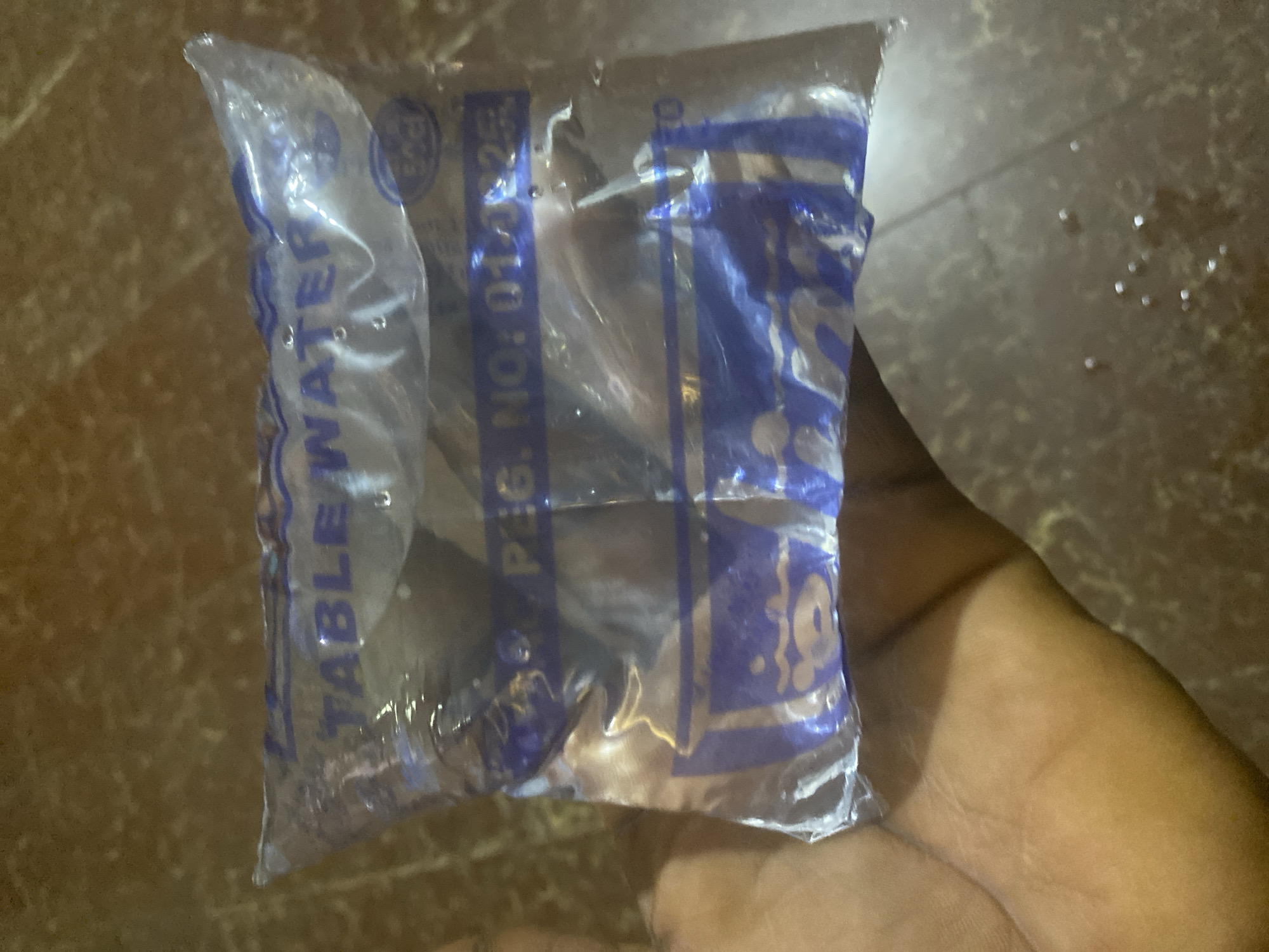 The image depicts a human hand holding a clear plastic sachet containing water. The sachet features blue text and graphics. Prominently visible text on the top band reads "TABLE WATER". Below this, another blue band displays "PEG. NO: 014925", with the last two digits partially obscured. A blue graphic, possibly a stylized water droplet or container, is printed on the lower portion of the sachet. The hand, exhibiting a dark skin tone, is seen from the palm and finger side, supporting the sachet from beneath. The background consists of a dark, mottled surface, likely a floor or table, upon which several small, reflective water droplets are scattered. The setting appears to be indoors with ambient lighting. While the image shows a product commonly found in Jos, Nigeria, specific visual identifiers for the city or an exact location within it are not present in the photograph.