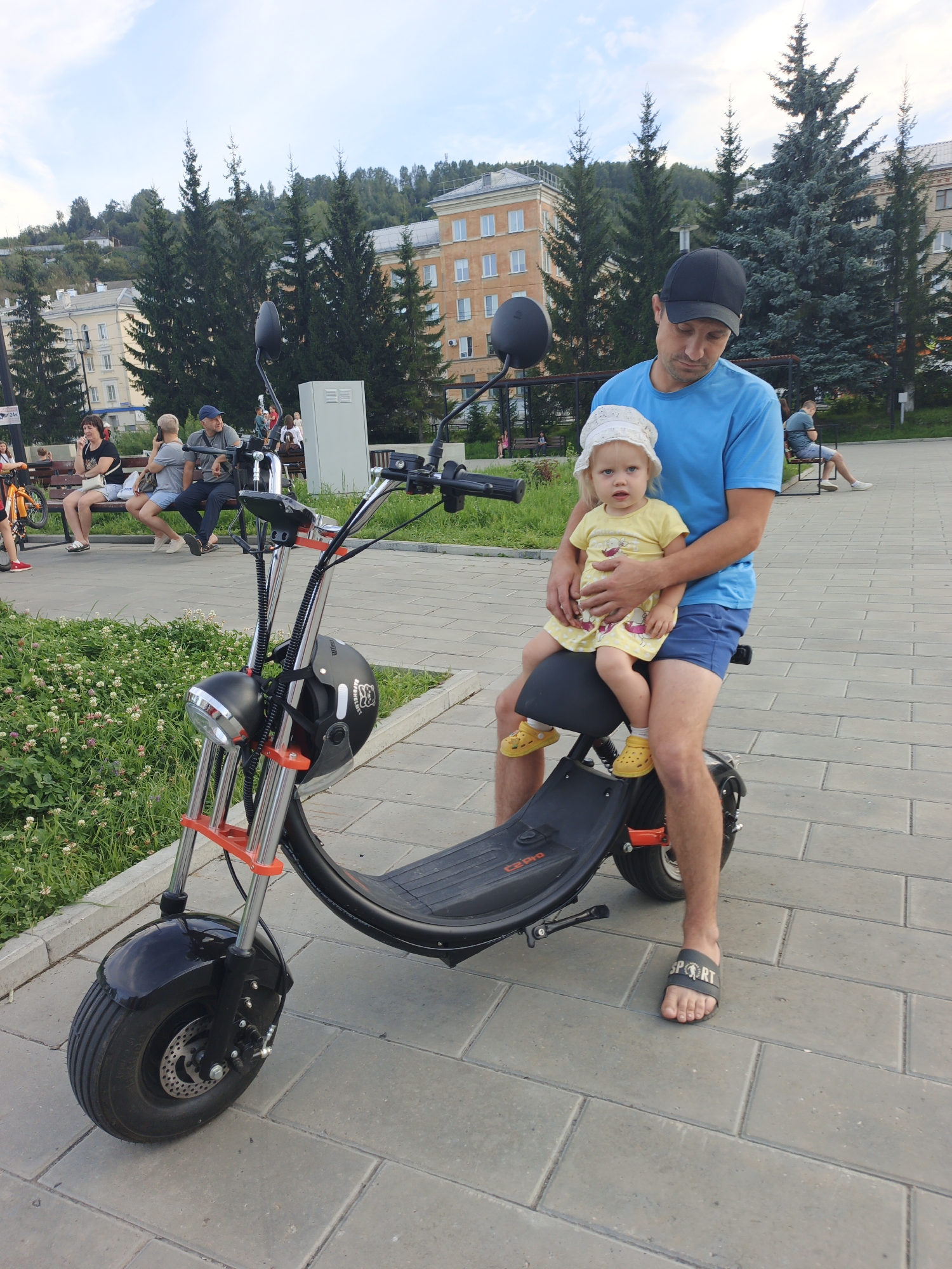 A man in a blue t-shirt and blue shorts is sitting on an electric scooter, holding a young child in a yellow dress and a white sun hat. The child is looking at the camera. The man is wearing a black baseball cap and black slide sandals with "SPORT" written on them. The scooter is black with orange accents and a black helmet with a white logo is attached to the handlebars. In the background, there are other people sitting on a bench, trees, and buildings. The scene appears to be in an urban park or pedestrian area with paved walkways and grass. The location is Zlatoust, Russia.