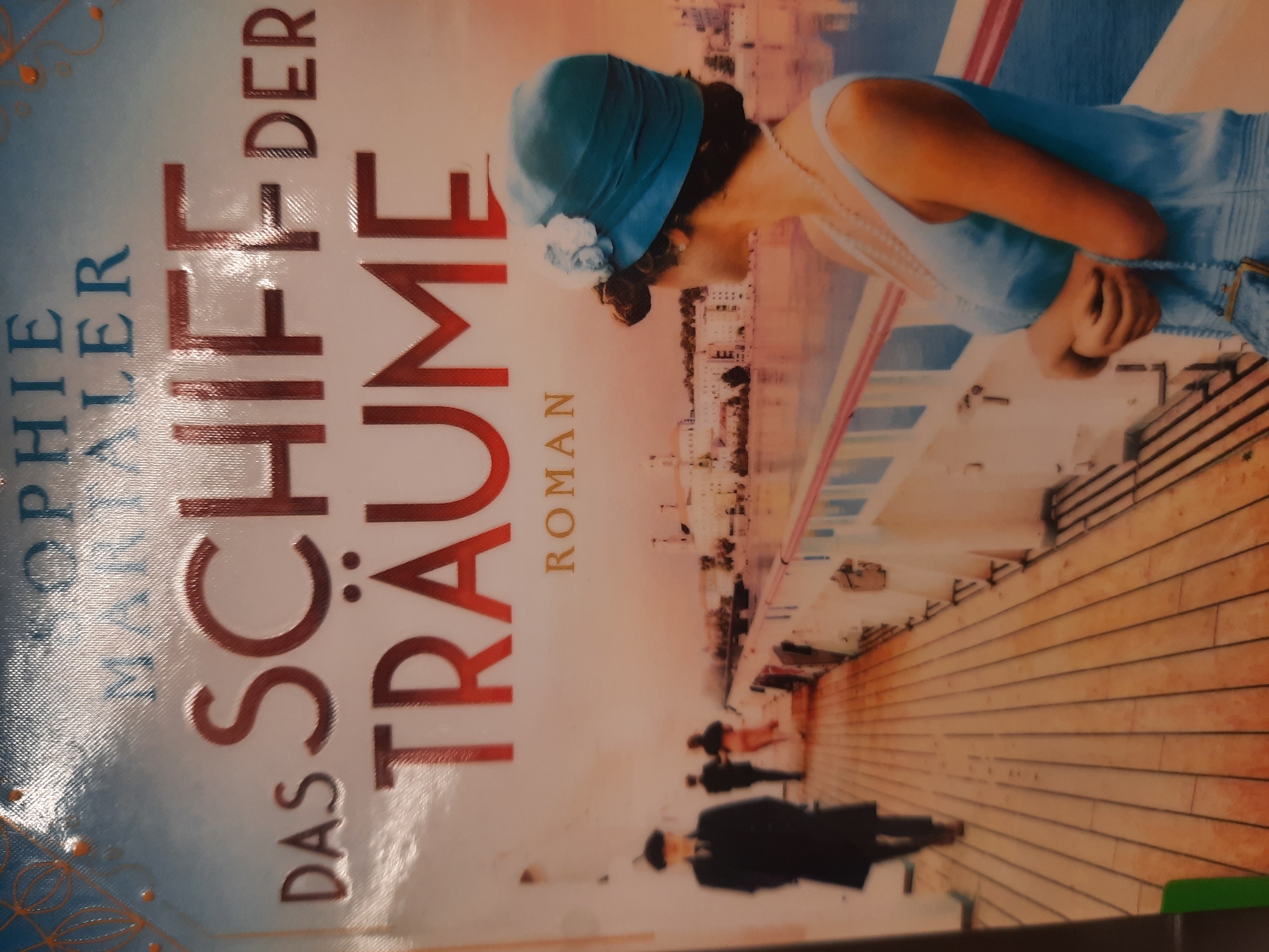 The image is a close-up of a book cover with the title "DAS SCHIFF DER TRÄUME" and the author's name "SOPHIE MARTALER". The cover depicts a scene on the deck of a ship, with several people in the background. In the foreground, a woman in a blue dress and hat looks out at a cityscape across the water. The cover art is vibrant and appears to be a painting or illustration. There are no indications of the city of Rosenheim, Germany within the image itself.