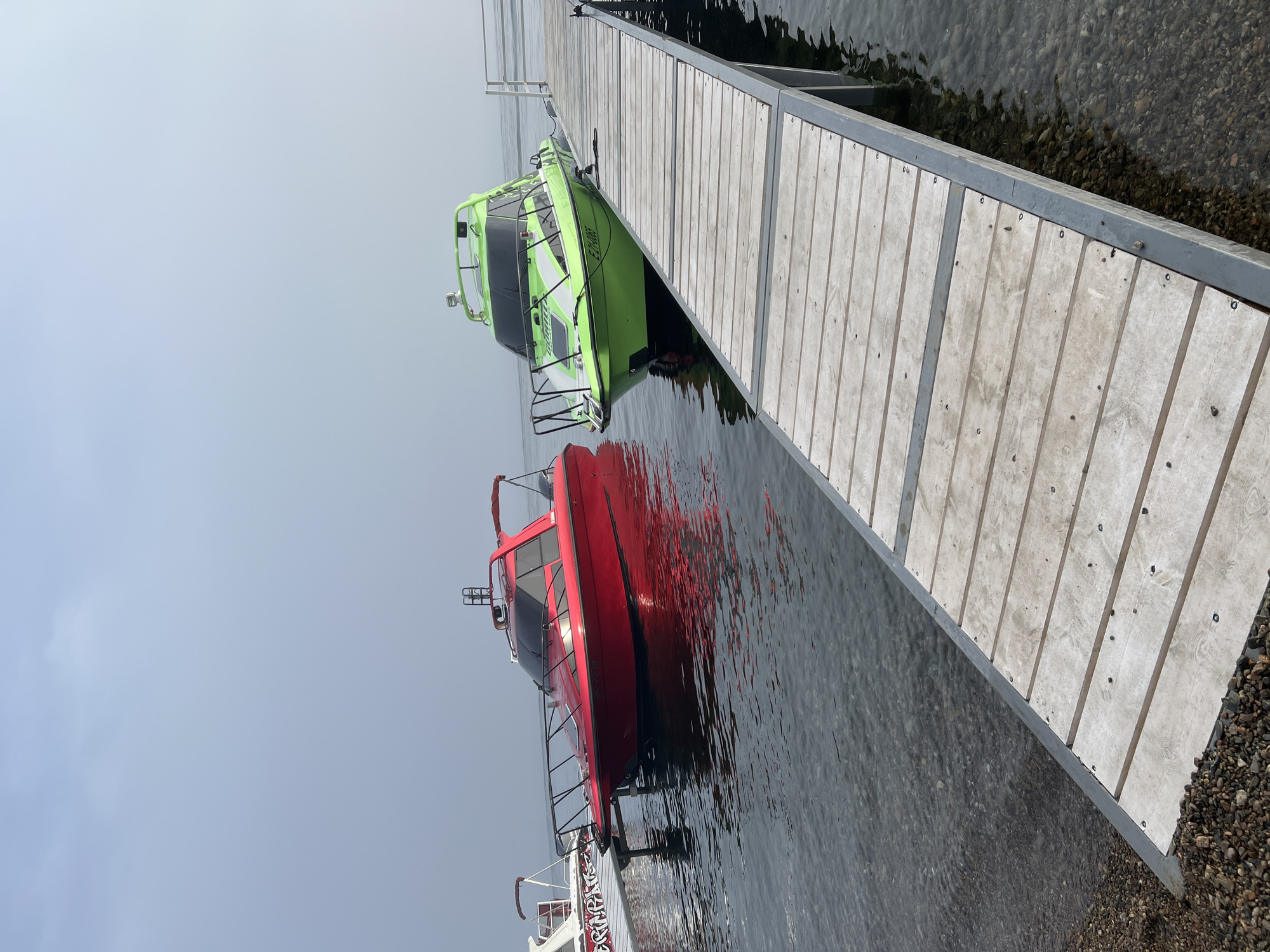 Two speedboats, one red and one lime green, are docked at a wooden pier on a body of water. The red boat is on the left, and the green boat is on the right. The pier extends diagonally from the bottom right corner of the frame towards the boats. The water is clear and shows the rocky bottom. The sky is overcast and hazy. The scene appears to be a calm day at a dock in Listvyanka, Russia, a settlement located on the southwestern shore of Lake Baikal.