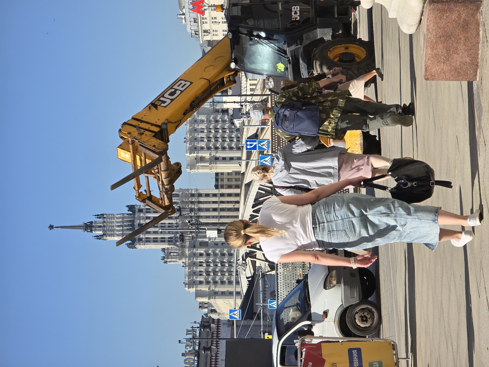 The image shows a street scene in Moscow, Russia, taken during the day under clear blue skies. A yellow JCB construction vehicle is positioned on the right side of the frame, partially obstructing the view of a white and beige Stalinist skyscraper in the background. Several pedestrians are present, including a woman in a white t-shirt and blue denim skirt, standing nearest to a white compact van. Another person is wearing a grey shirt and pink shorts, and a third person is wearing camouflage clothing. Pedestrian crossing signs are visible near the van.
