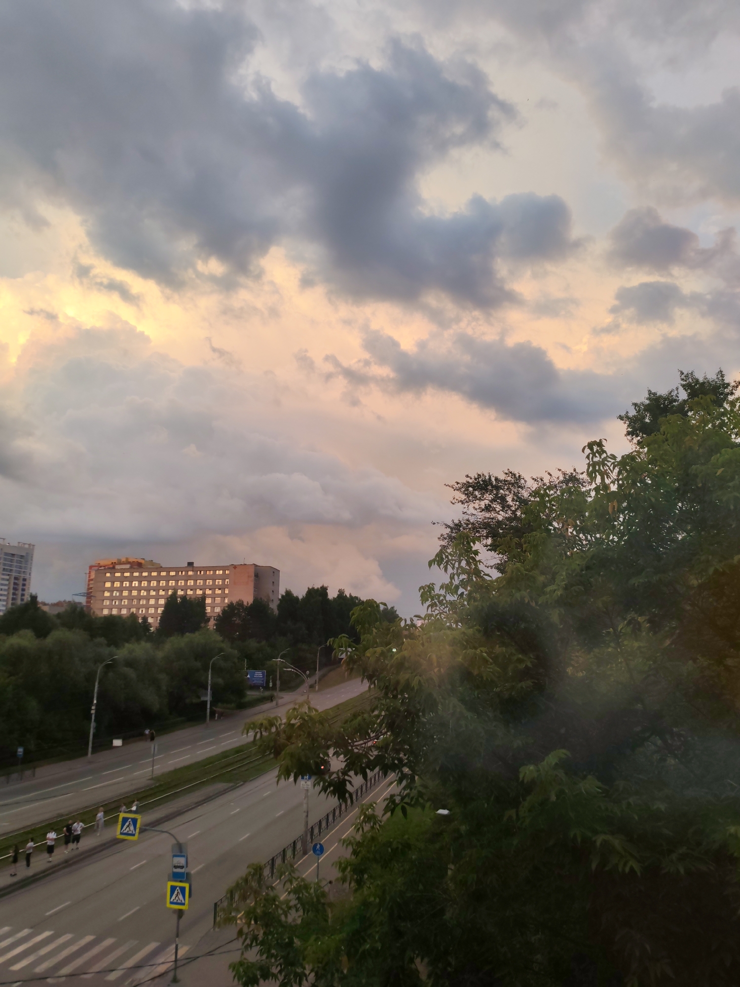 The image depicts a street scene in Izhevsk, Russia, during what appears to be late afternoon or early evening. The sky is filled with dramatic, cloudy formations, with patches of light peeking through, suggesting the sun is setting or has recently set, casting a warm glow on the clouds. In the background, a multi-story building with numerous windows is visible, surrounded by trees. A street runs through the midground, with several lanes of traffic. Pedestrians are visible walking on the sidewalk alongside the street, and there are traffic signs and lampposts. Lush green trees dominate the foreground and right side of the image, with their foliage partially obscuring the view. The overall impression is a city landscape with natural elements and man-made structures under a dynamic sky.