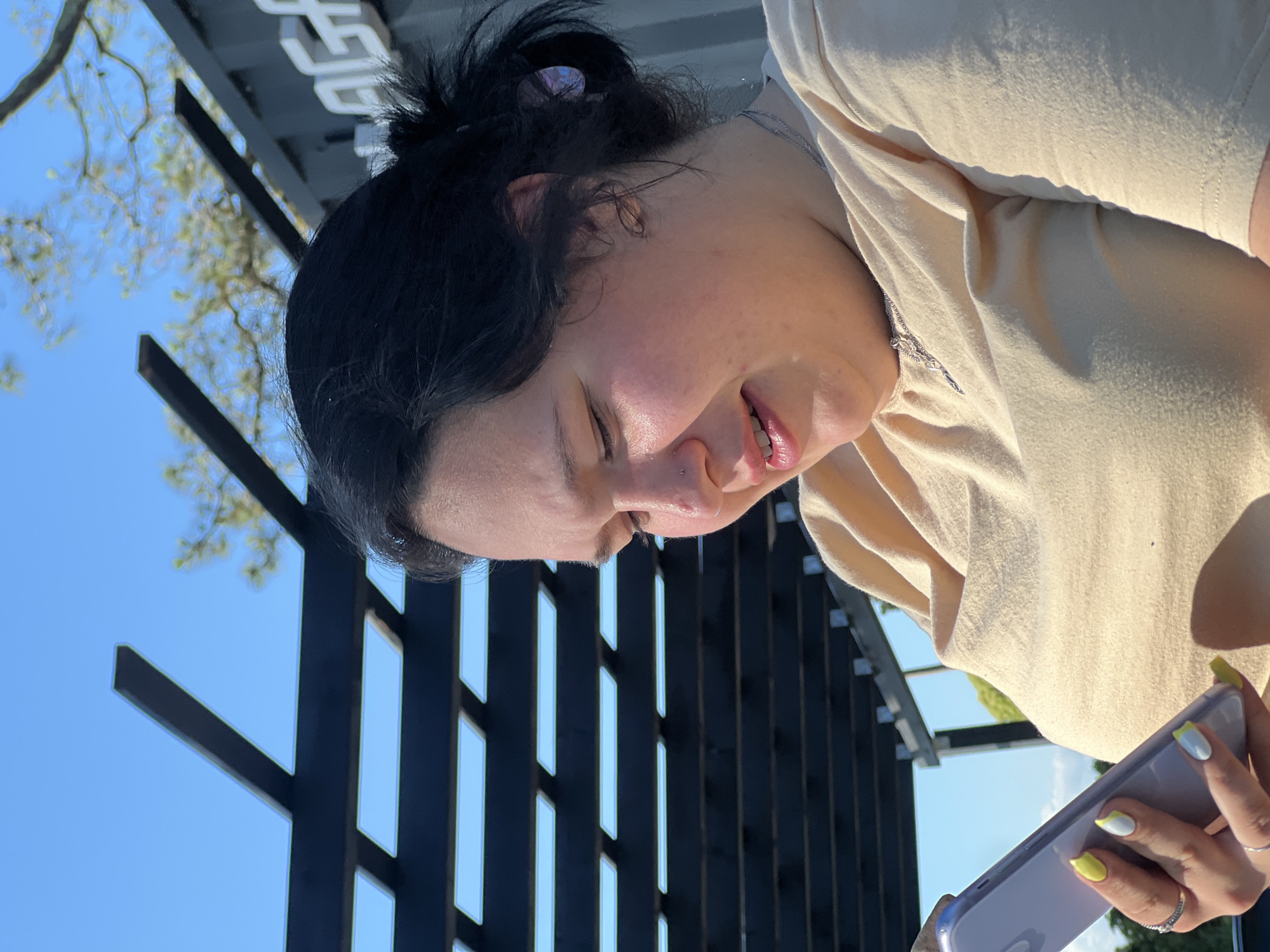 A medium shot shows a young woman with dark hair pulled back in a messy bun and fair skin looking down at her phone. She is wearing a light yellow t-shirt and a silver necklace. She has painted nails in white with yellow accents, and rings on her fingers. She is outdoors, with a dark wooden pergola structure in the background and a bright blue sky above. Trees are visible in the distance, along with part of a building. The scene appears to be in a public outdoor space in the city of Ochamchire, Abkhazia.