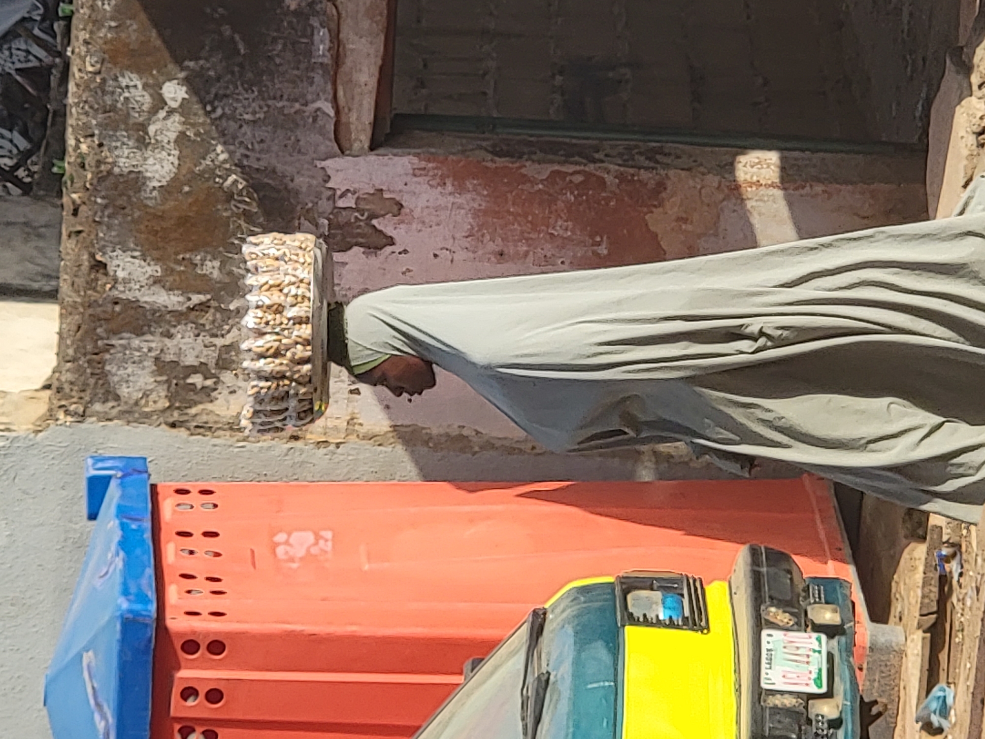 The image captures a man in Maiburiji, Nigeria, likely a Muslim, wearing a taqiyah cap and draped in a grey, draped cloth, possibly a prayer garment. The man is outside, adjacent to a dilapidated building with crumbling white-painted walls and a worn, reddish wall section above. An orange and blue truck is parked nearby, its front end featuring yellow panels and a partially visible license plate with illegible text. The overall scene depicts an everyday moment in a weathered urban environment.