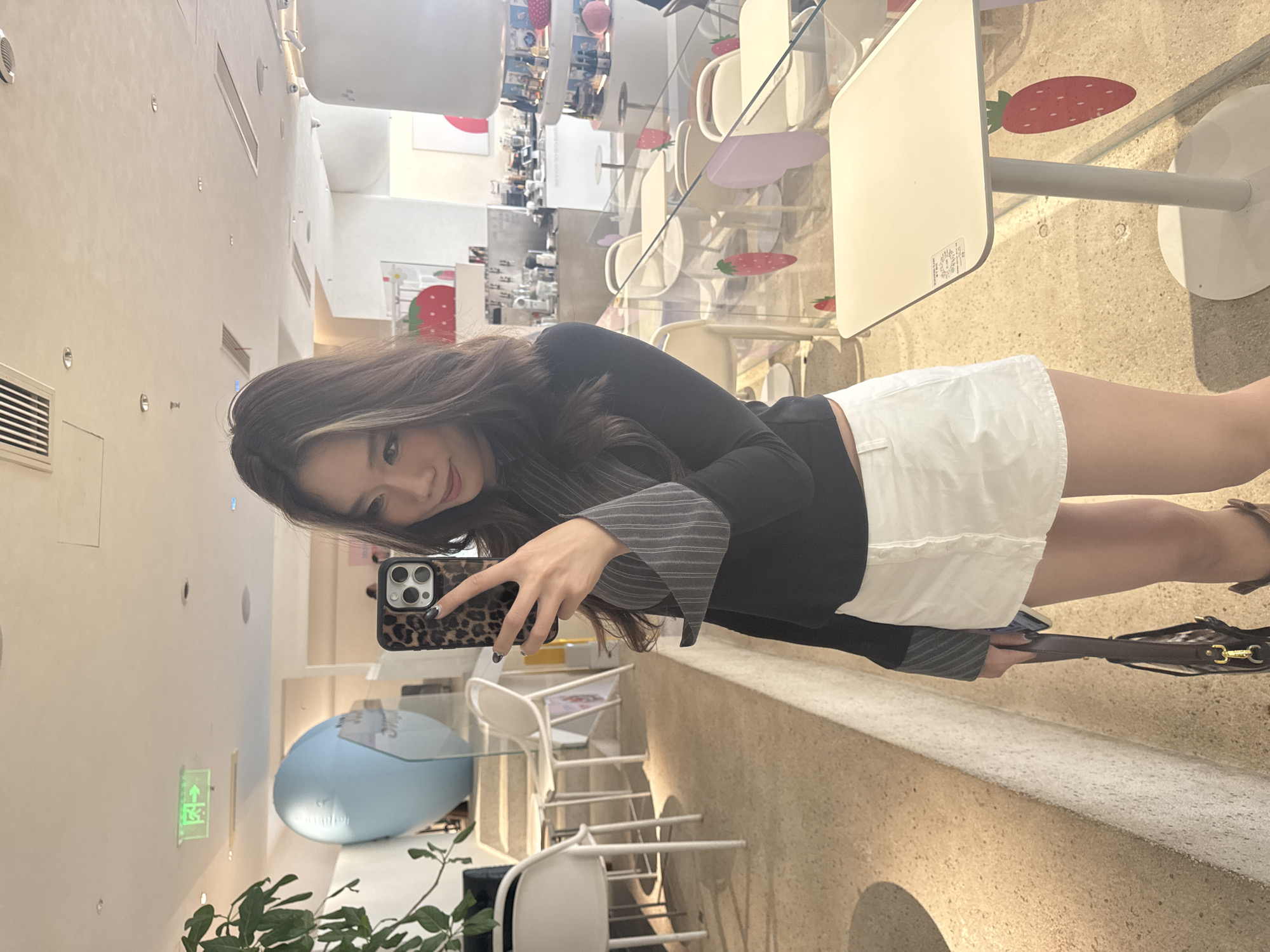 A woman stands in a cafe in 江北城街道, China, taking a selfie. She holds a leopard-print phone in her left hand. The cafe is decorated with strawberry-themed elements. Tables with glass tops and white chairs are visible. A blue, dome-shaped object is also in the background. A plant is in the foreground.