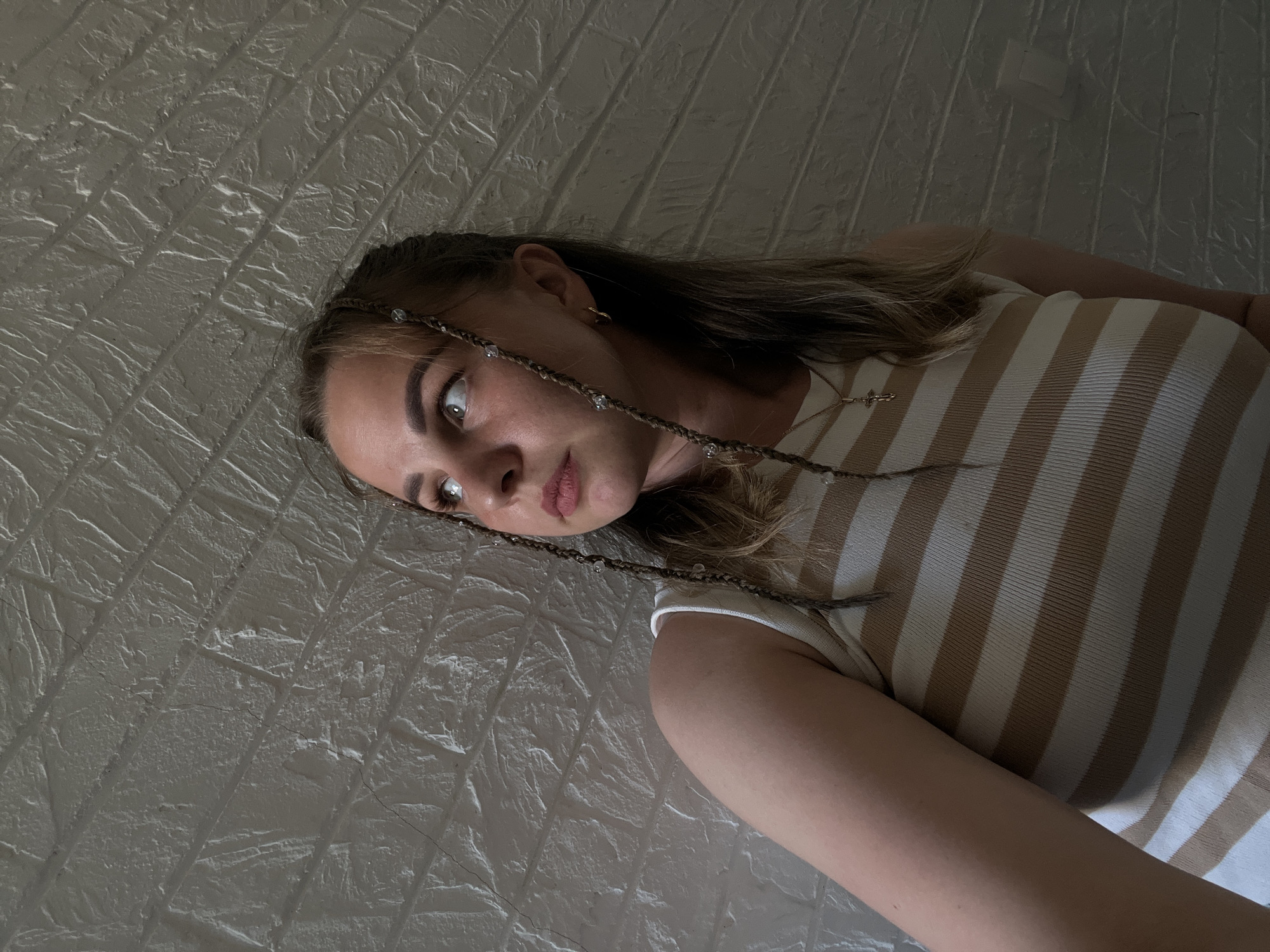 A young woman lies on a white textured surface. She is wearing a striped beige and white shirt. Her hair is light brown with thin braids adorned with small, clear beads. She is looking to the left. The surface appears to be a brick-patterned wall or floor. The scene is dimly lit. There is no information indicating the location is Izhevsk, Russia.