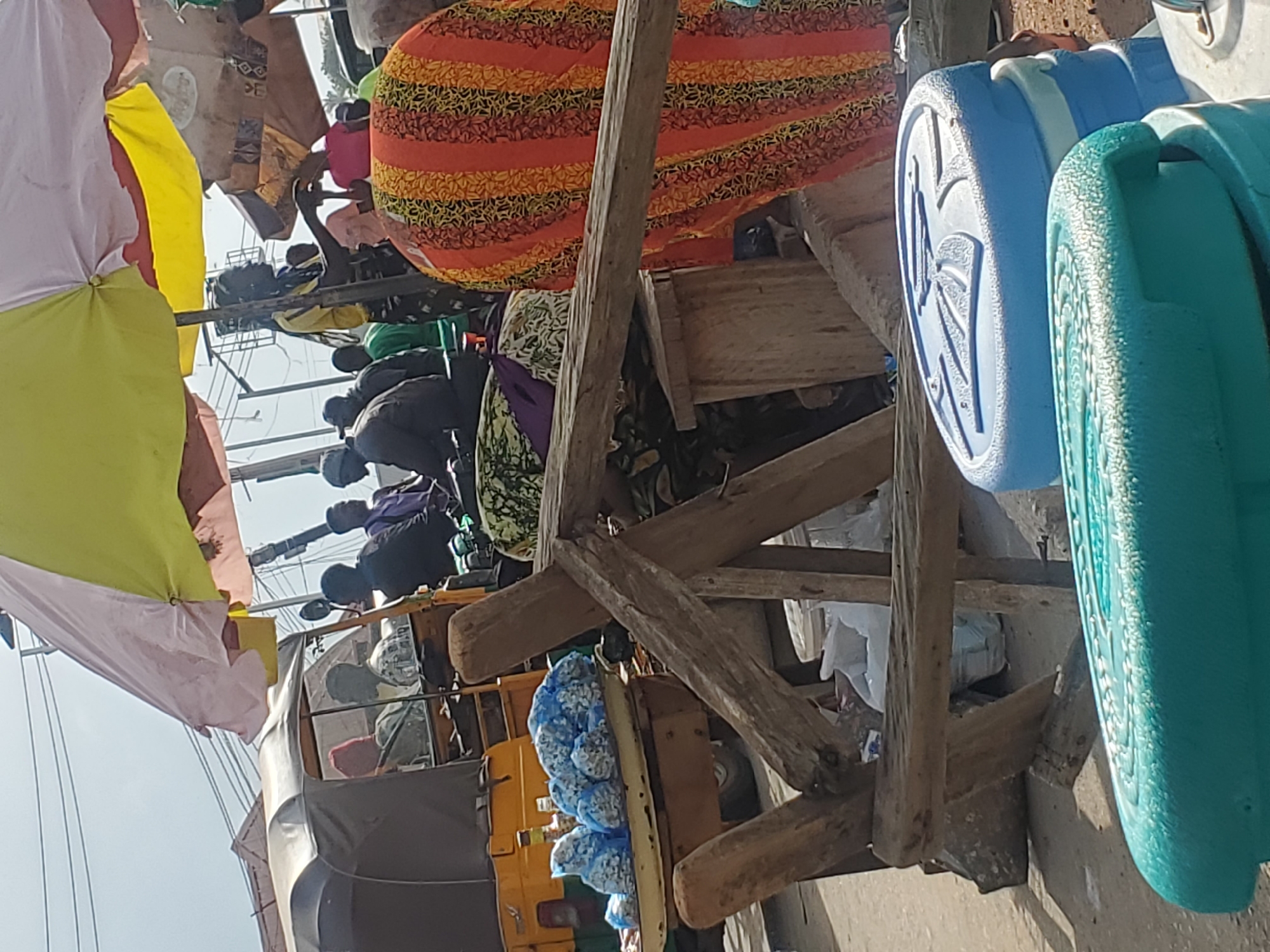 The image depicts a street scene, likely a market, in Gwong, Nigeria. The foreground features two large, stacked plastic containers, one turquoise and the other blue, with patterned lids. Behind these, a wooden structure, possibly a makeshift stall or cart, is visible, made of rough-hewn logs. To the left, a yellow auto-rickshaw, also known as a "Keke Napep," is parked. On a tray in front of the auto-rickshaw, numerous small items are packaged in blue plastic bags. The middle ground shows several people, some in the auto-rickshaw, and others standing or walking. One person, wearing a bright orange and yellow patterned garment, is prominent in the middle right of the frame. A large, multi-colored umbrella (yellow, pink, and white) provides shade over part of the scene. The background includes overhead wires and more indistinct figures and structures, suggesting an urban environment. The overall atmosphere is one of a busy market or transportation hub.