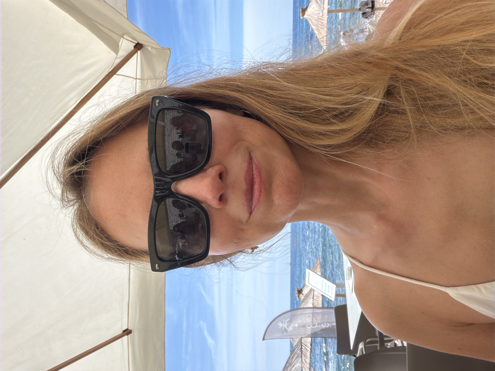 A blonde woman with sunglasses is taking a selfie at a beachside cafe in Budva, Montenegro. She is wearing a white swimsuit and her hair is blowing in the wind. In the background, the sea and sky are visible, along with straw umbrellas and beach chairs. The reflection in her sunglasses shows other patrons at the cafe.