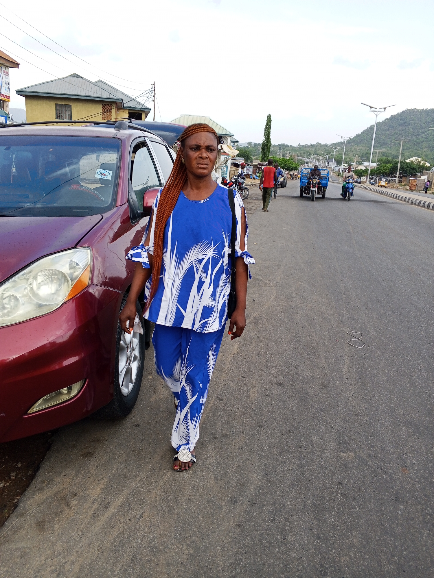 The image portrays a street scene in Zango-ndaji, Nigeria, under an overcast or hazy sky, suggesting late afternoon or early evening. In the foreground, a woman with brown braided hair walks on the asphalt road, positioned to the right of a maroon minivan. She wears a matching blue and white patterned top, featuring tie details on the sleeves, and coordinating pants. Her sandals are adorned with a circular silver embellishment, and she carries a small black bag slung over her shoulder. Her expression appears neutral.
The minivan, identified as a "TOYOTA" by a sticker on its windshield, also bears another sticker with "JNSC". The street continues into the midground, where several people and vehicles are visible. A man walks in the distance, and two other men ride motorcycles; one operates a motorcycle with a blue cargo attachment, typical of a tricycle.
The roadside features utility poles and streetlights, some of which are equipped with solar panels. The road itself has a curb marked with black and white stripes. In the background, various buildings line the street, including a prominent light yellow house with a green roof. Distant green hills define the horizon, indicating a somewhat hilly or mountainous landscape. A small, bent metal object lies on the road surface in the midground. The scene captures everyday activity on a busy road.