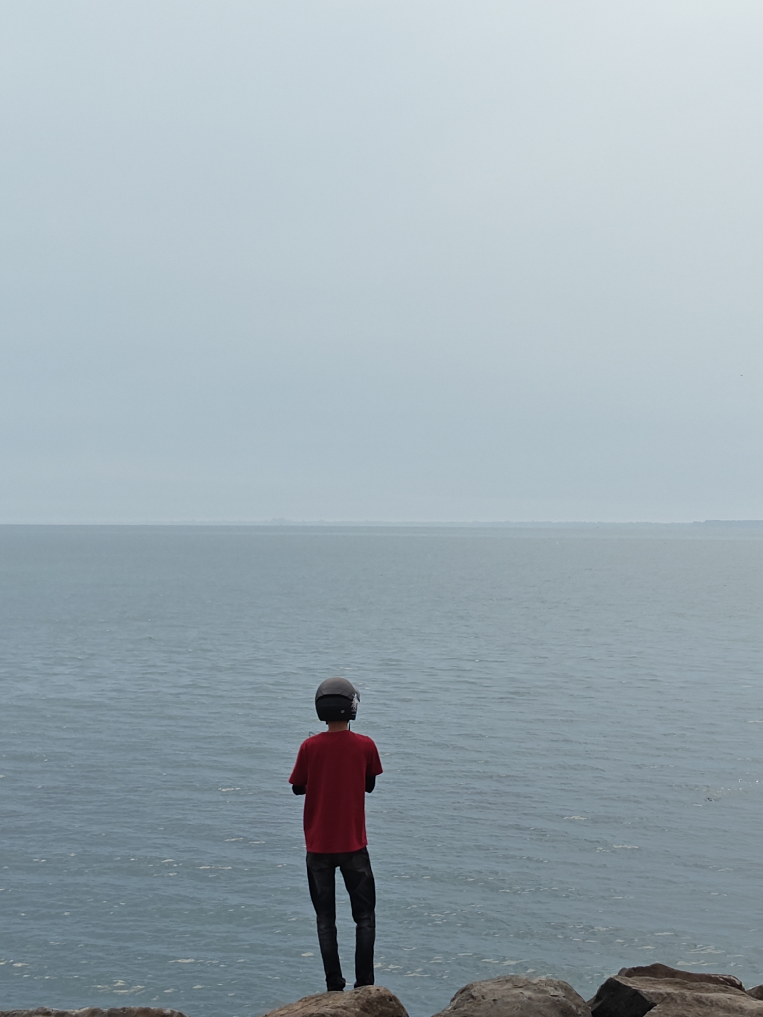 The image depicts a solitary figure standing on a rocky shore, looking out over a calm, expansive sea under an overcast sky. The person, viewed from behind, wears a red short-sleeved t-shirt, dark trousers, and a dark grey or black helmet. Their arms are crossed behind their back, suggesting a posture of observation or contemplation.
The scene is dominated by the vast body of water, which occupies the middle ground of the frame, stretching towards a distant, hazy horizon line. The water's surface shows subtle ripples, indicating calm to moderate conditions. Above the horizon, the sky is uniformly light grey, overcast, and diffused, casting soft, shadowless light over the entire landscape.
In the immediate foreground, large, dark rocks provide a sturdy platform for the figure. There are no other people or significant objects visible. The overall mood of the image is serene and perhaps contemplative, highlighting the individual's presence against the grandeur of nature. No text is visible. Based on the provided context, the setting is a coastal area in Lhokseumawe, Indonesia.
