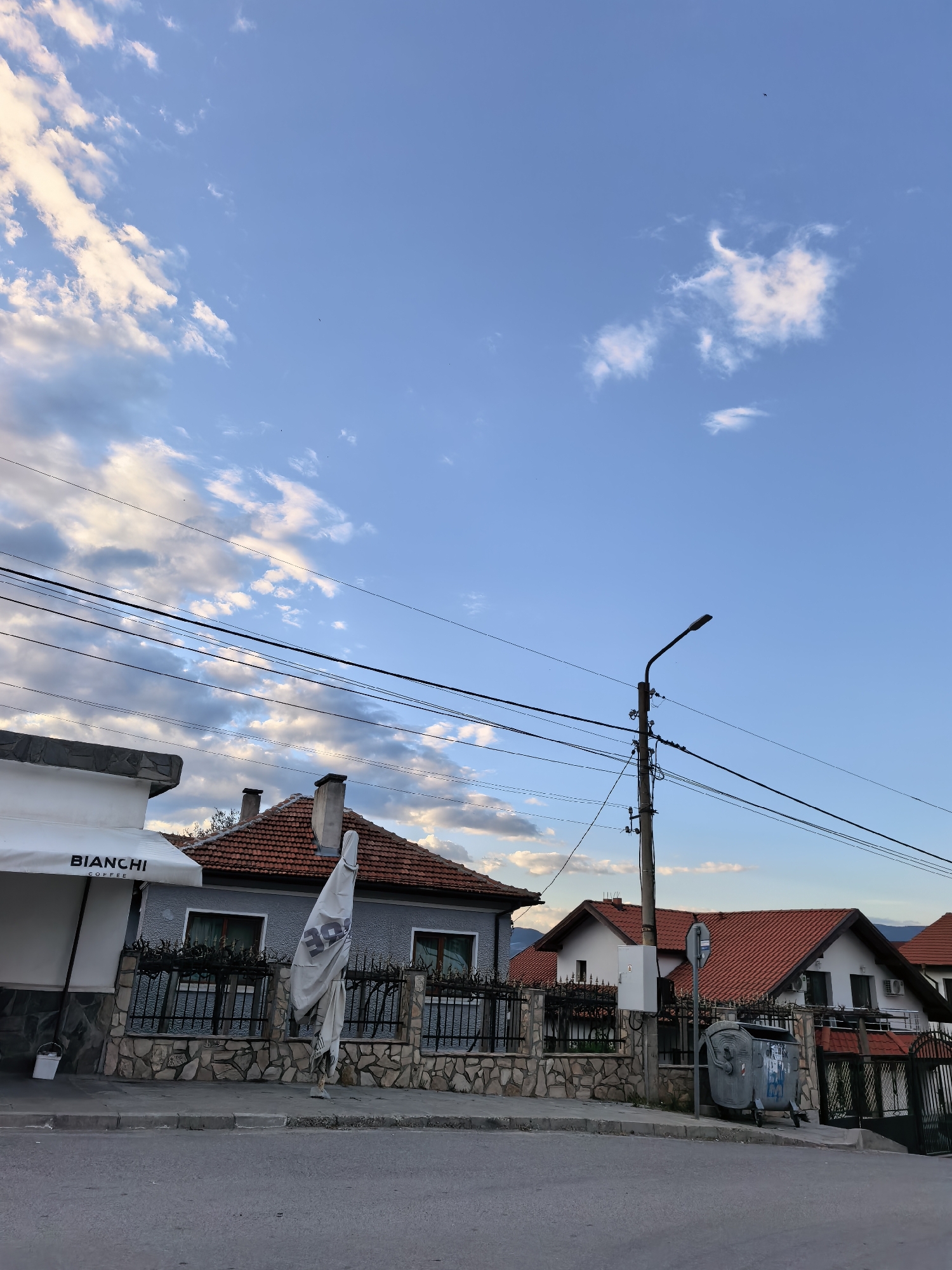 The image captures a street scene in an urban environment. The sky is predominantly blue with scattered clouds. Visible buildings with red-tiled roofs and grey exteriors line the street. A Bianchi Coffee establishment with a white awning is visible. A utility pole with numerous wires is present. Refuse containers are positioned by a fence. There are no people visible in the scene. The exact location in the city of Unknown, Unknown, cannot be determined from the given image.