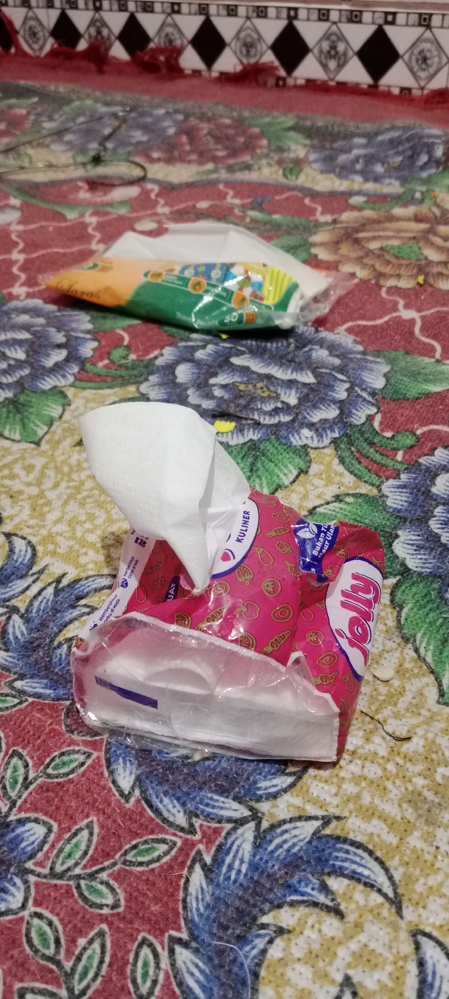 A packet of pink "Jolly" brand wet wipes is open with a few wipes protruding from it, placed on a floral-patterned tablecloth. Another partially visible packet of wet wipes, with an orange and green design, is positioned behind the pink packet. The background shows a part of a patterned border with red and black diamond shapes. The scene appears to be indoors, possibly in a home in Soko, Indonesia.