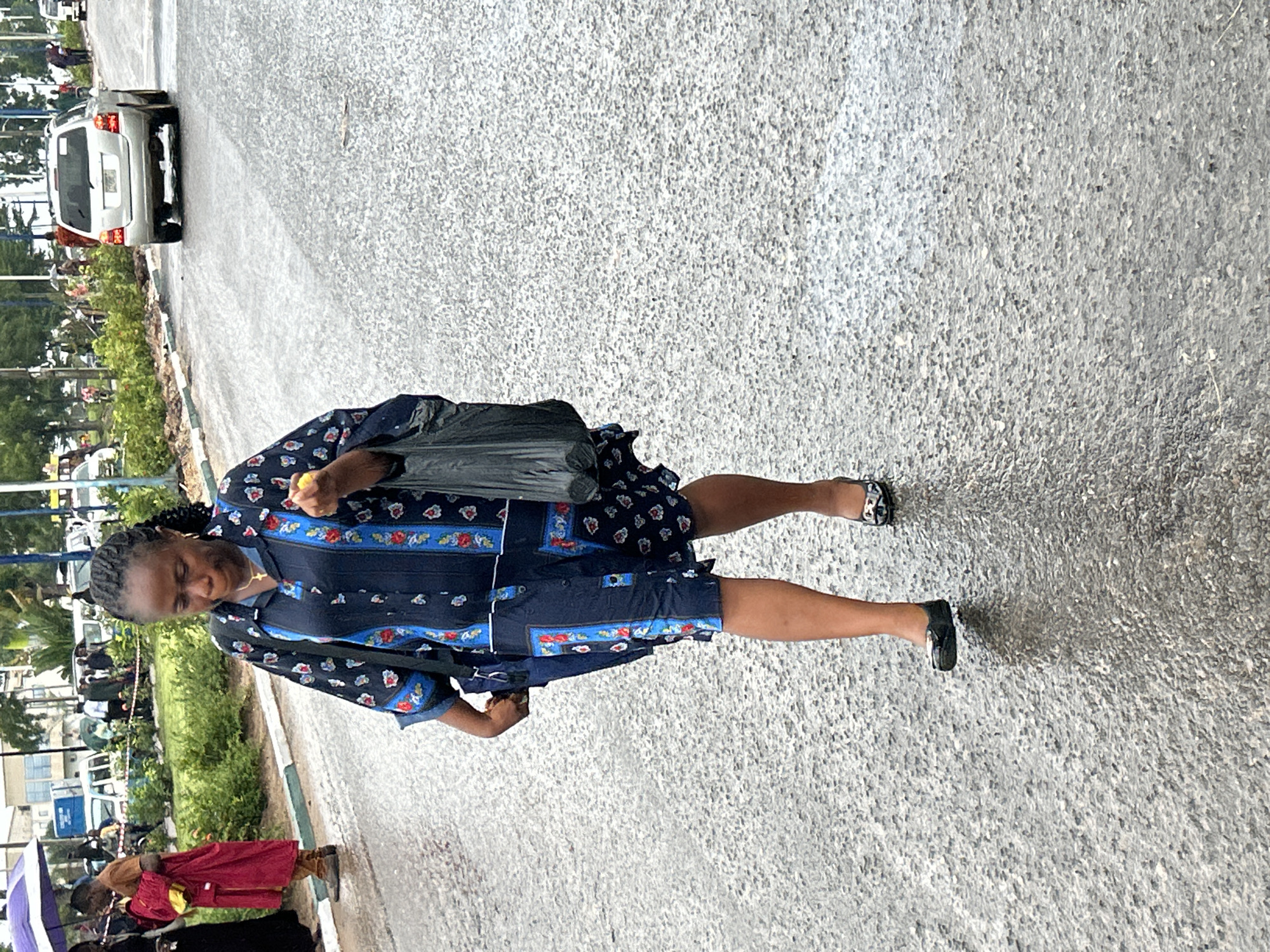 A woman with dark skin and braided hair walks on a wet paved road in Ihiagwa, Nigeria. She is dressed in a navy blue, collared, button-down dress featuring red, white, and blue floral patterns, accented with vertical blue patterned panels. A gold cross pendant hangs from her neck. She carries a black shoulder bag, a black plastic bag in her left hand, and a yellow object in her right hand. Her footwear consists of black flat shoes, one adorned with a metallic embellishment. A clear shadow extends behind her on the wet asphalt.
To the left, behind a curb and a landscaped strip of green bushes, several individuals are visible. One person wears a long reddish-brown garment and carries a yellow bag, standing near a woman partially sheltered by a purple umbrella. Other indistinct figures and vehicles, including a white van, are present further in the background.
To the right, a silver SUV, consistent with a Toyota Highlander, is driving away from the viewer on the wet road. Its license plate is partially visible. Additional vehicles and structures line the road in the distance. The overall environment indicates recent rainfall, with a damp road surface and green foliage lining the roadside.