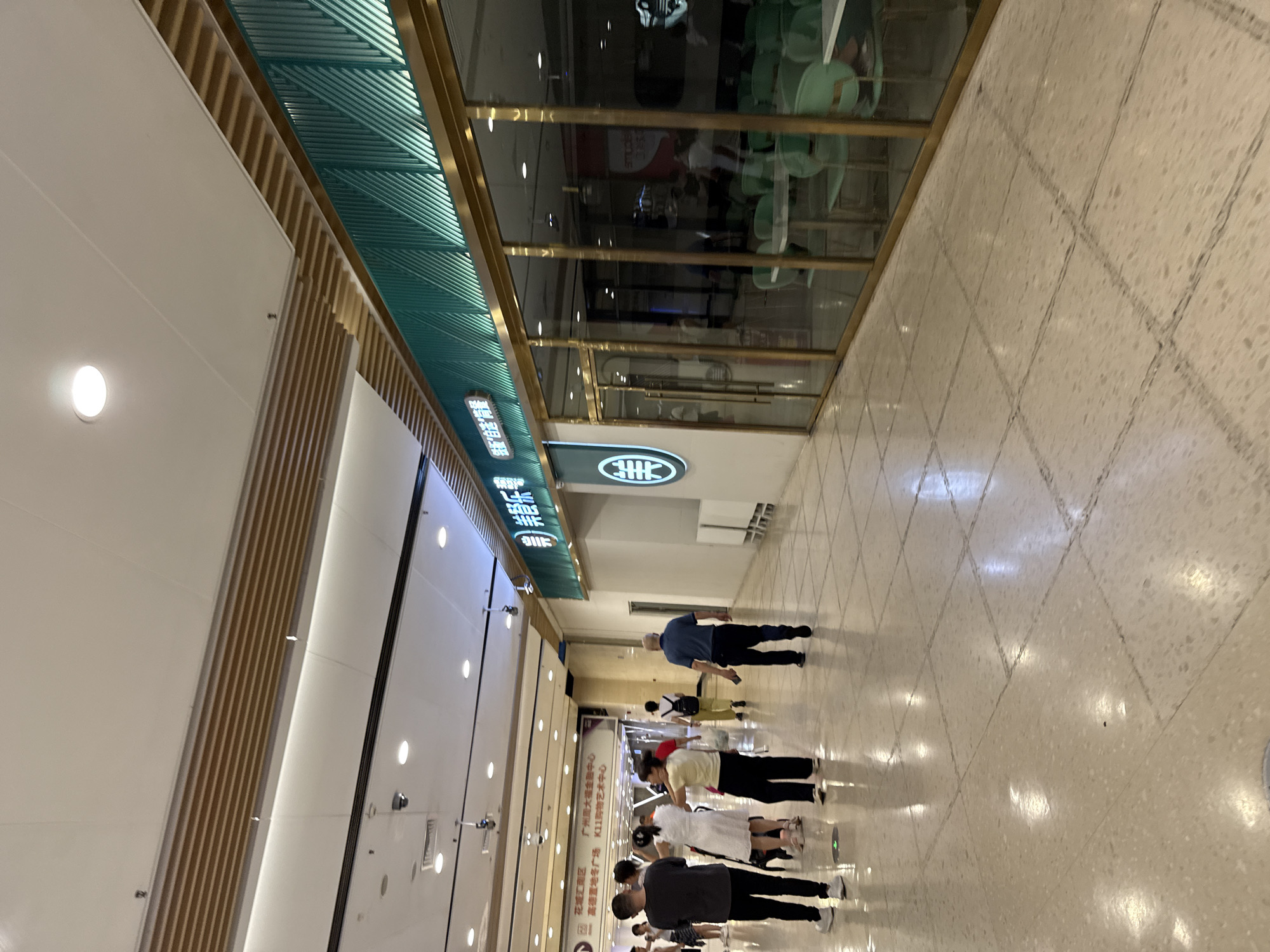 The image captures an interior view of a shopping mall, likely in Tianhe District, China, as indicated by the signage. The scene is well-lit with overhead lighting and shows a wide, polished floor reflecting the ambient light. In the foreground, several individuals are walking or standing, their backs mostly to the camera. To the right, a restaurant with a modern facade is visible. The restaurant features large glass windows, through which tables and light green chairs can be seen, suggesting it is open and operational. The restaurant's signage, written in Chinese characters with a prominent teal green border, is mounted above the entrance. The overall environment appears to be a commercial walkway within a large building, with architectural elements like suspended ceilings and lighting fixtures characteristic of a modern mall.