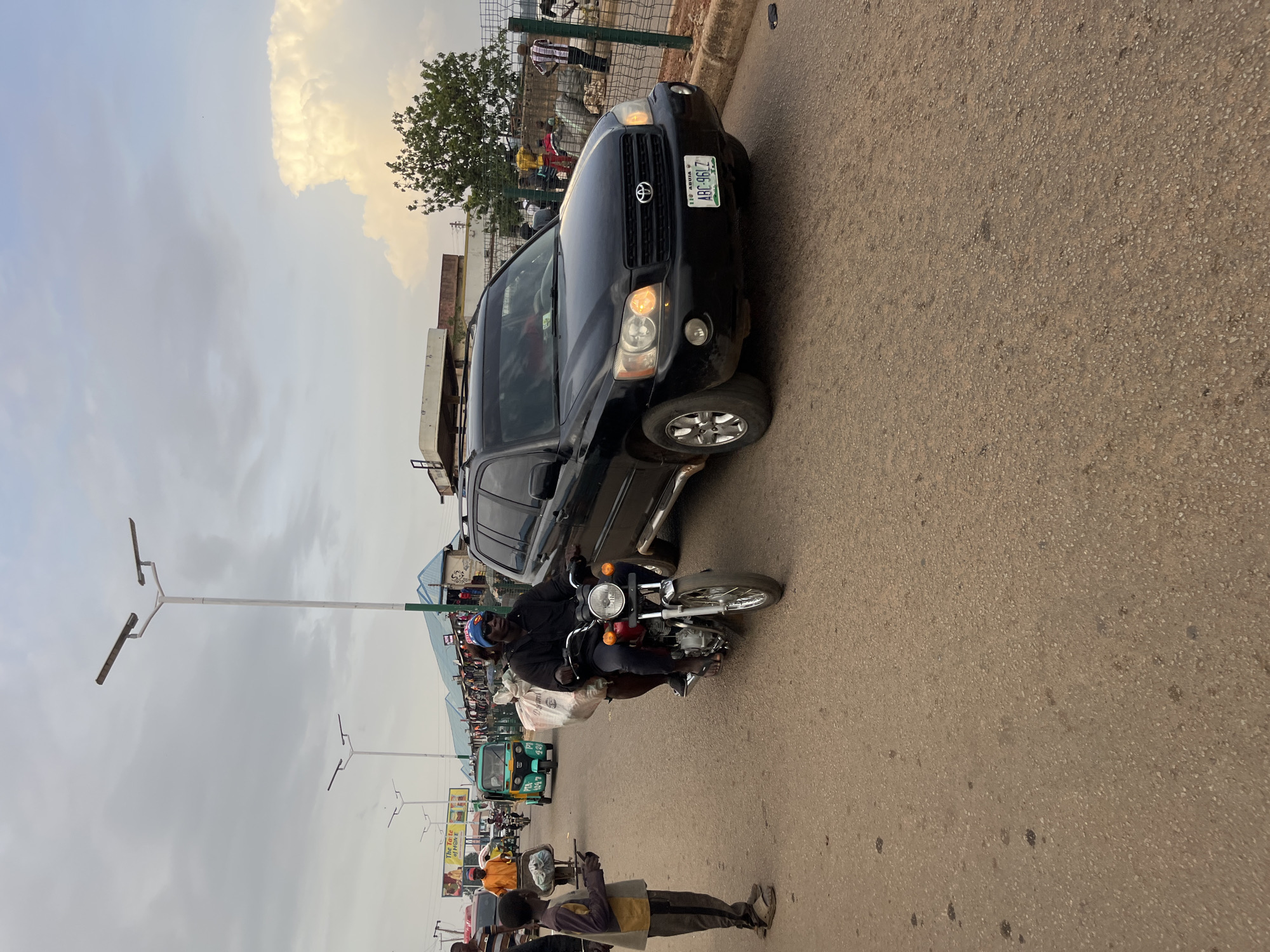 In a Nigerian city, possibly Bukuru, a street scene unfolds under a cloudy sky. A man wearing a blue beanie and sunglasses rides a red motorcycle, carrying a large white sack on the back. Beside him, a young boy in a grey and yellow shirt and dark pants stands on the side of the road. In the background, a black Toyota Highlander SUV drives forward, its headlights on. Other vehicles, including a green auto-rickshaw, are also present on the road. Market stalls with blue roofs line the street, and people mill about the area. The road is paved and appears to be well-trafficked.