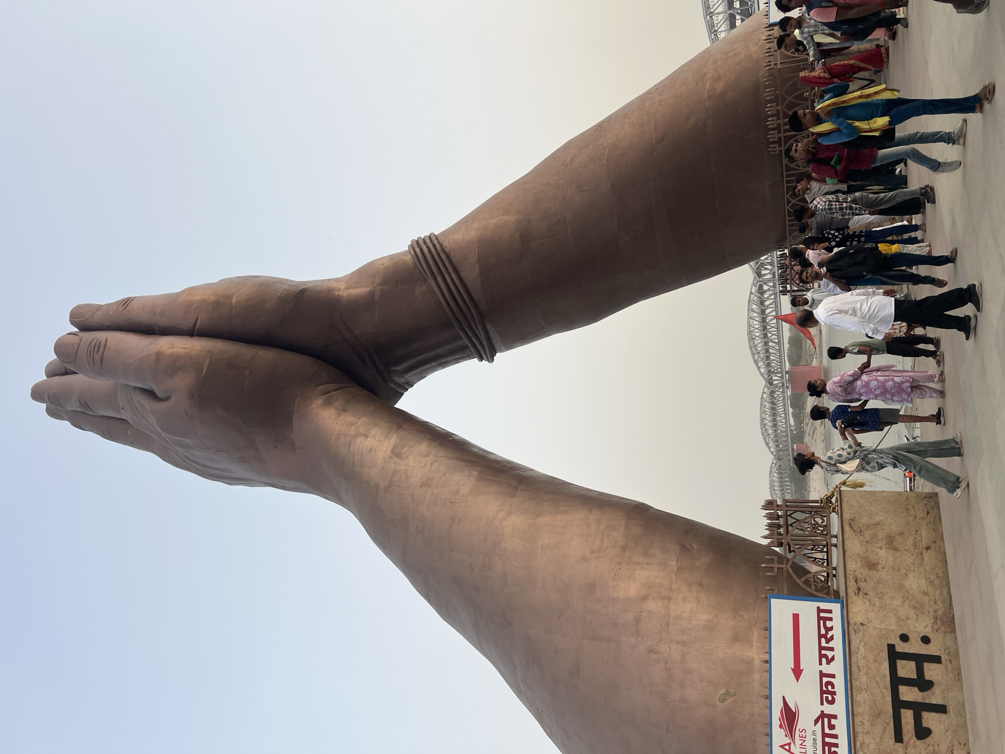 The image shows a large sculpture of two hands in a "namaste" gesture, located in Mughalsarai, India. The hands are bronze-colored. A thin bracelet adorns one wrist. Many people are standing in the background. A river appears between the sculpture and the background. A sign written in Hindi is visible at the base of the sculpture.