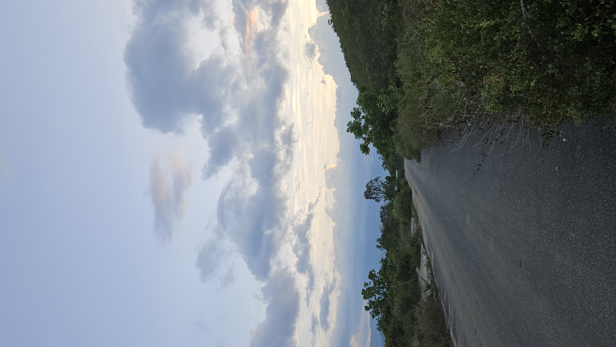 The image shows an asphalt road stretching into the distance, bordered by dense vegetation on one side and a cloudy sky on the other. The location is Cam Lam, Vietnam. No people are visible. The sky has a mix of blue and grey clouds. The dense vegetation on the road's side suggests a rural or less developed area.
