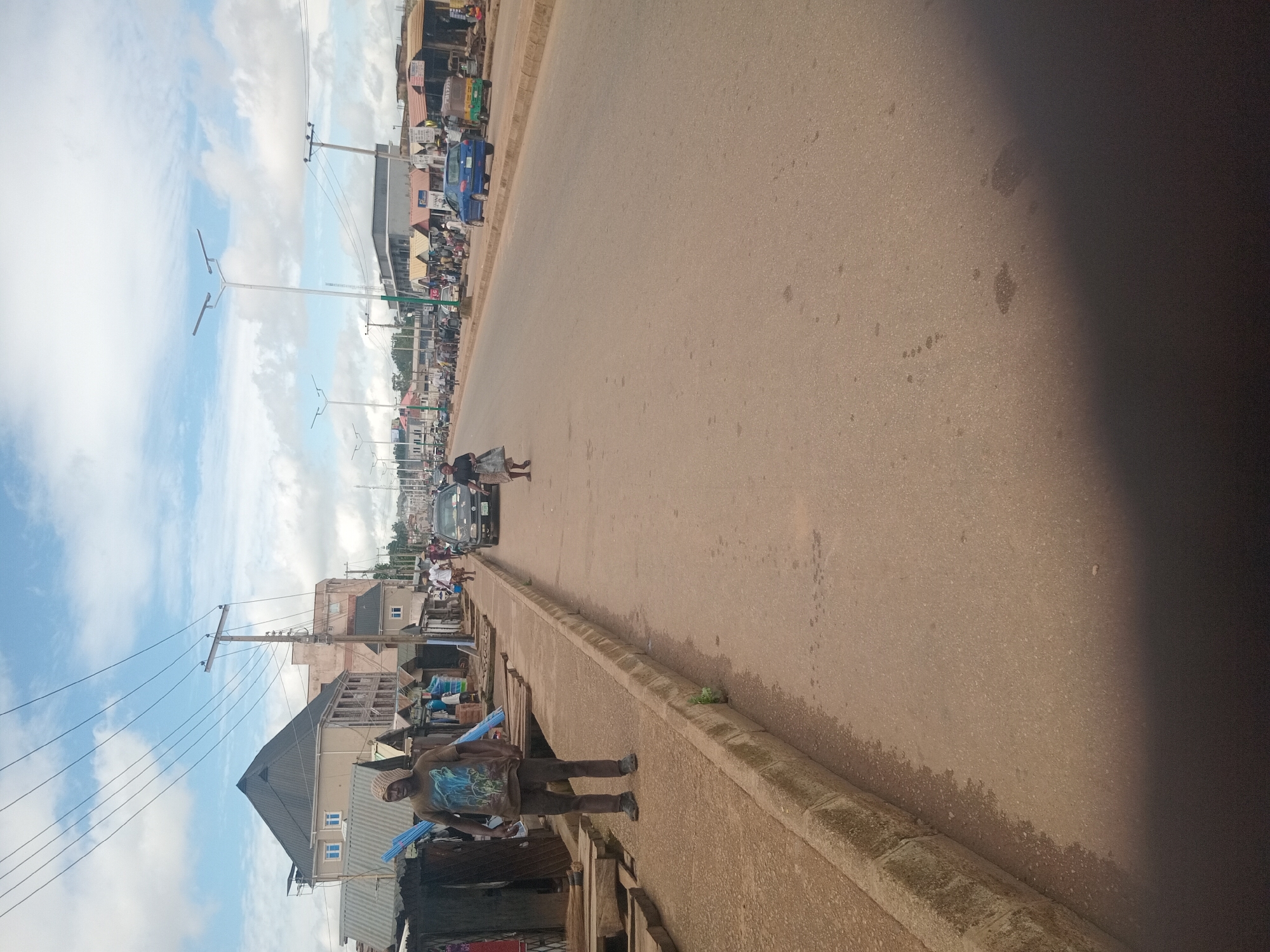 The image depicts a street scene in Bukuru, Nigeria. A man wearing a patterned t-shirt and dark pants stands on a concrete sidewalk next to a drainage ditch filled with dirt and debris. He is facing away from the camera, looking towards a row of market stalls. In the background, several market stalls are visible, along with buildings, streetlights, and a paved road. A few vehicles, including a blue car and a tricycle-like vehicle, are on the road. The sky is partly cloudy. The overall environment appears to be a busy commercial area.