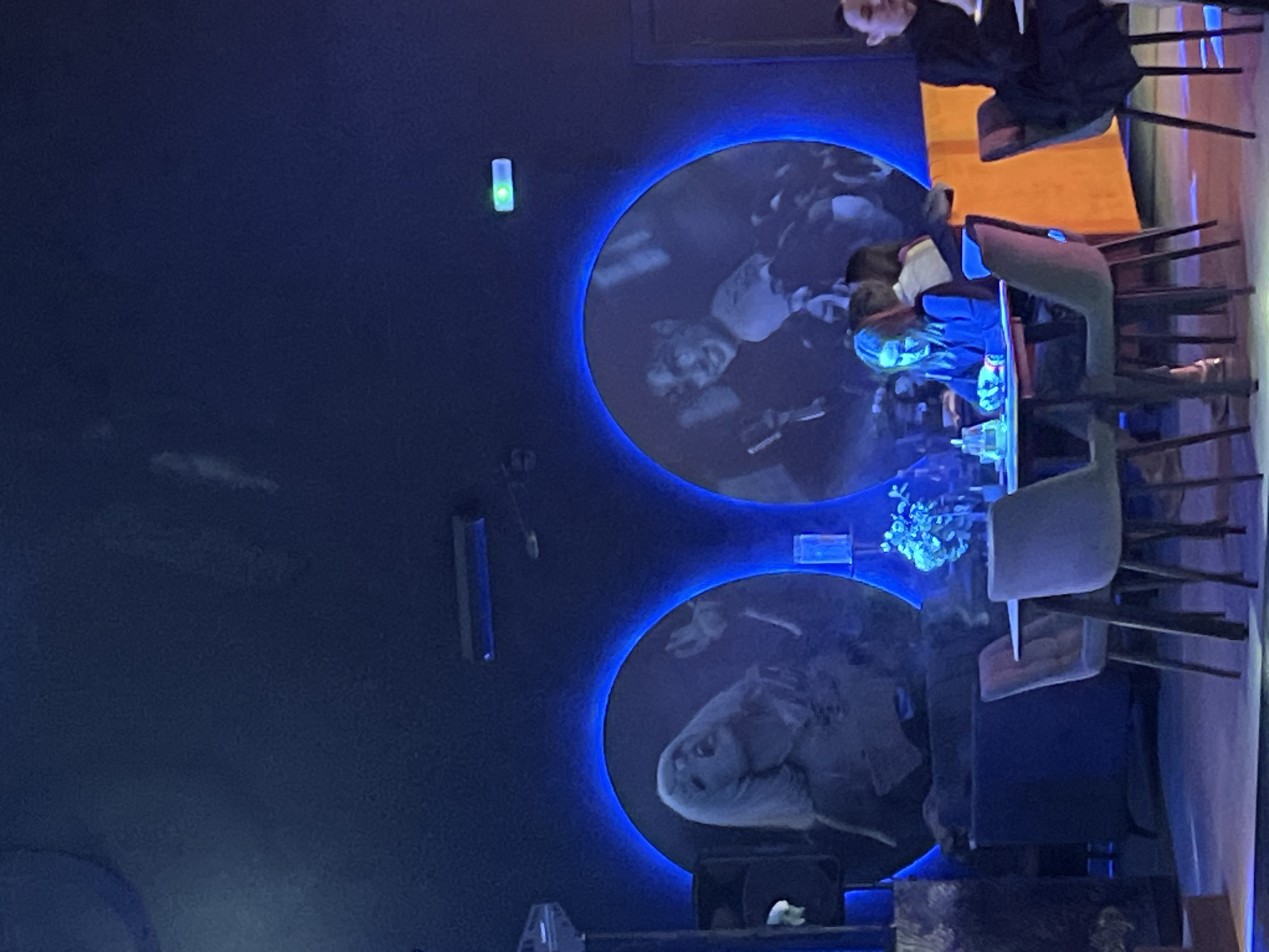 The image shows an interior view of a restaurant with a dark blue wall featuring two illuminated round portraits. Patrons are seated at tables, some of whom are eating or talking. The location is the Vремя cafe, in Tyumen, Russia.