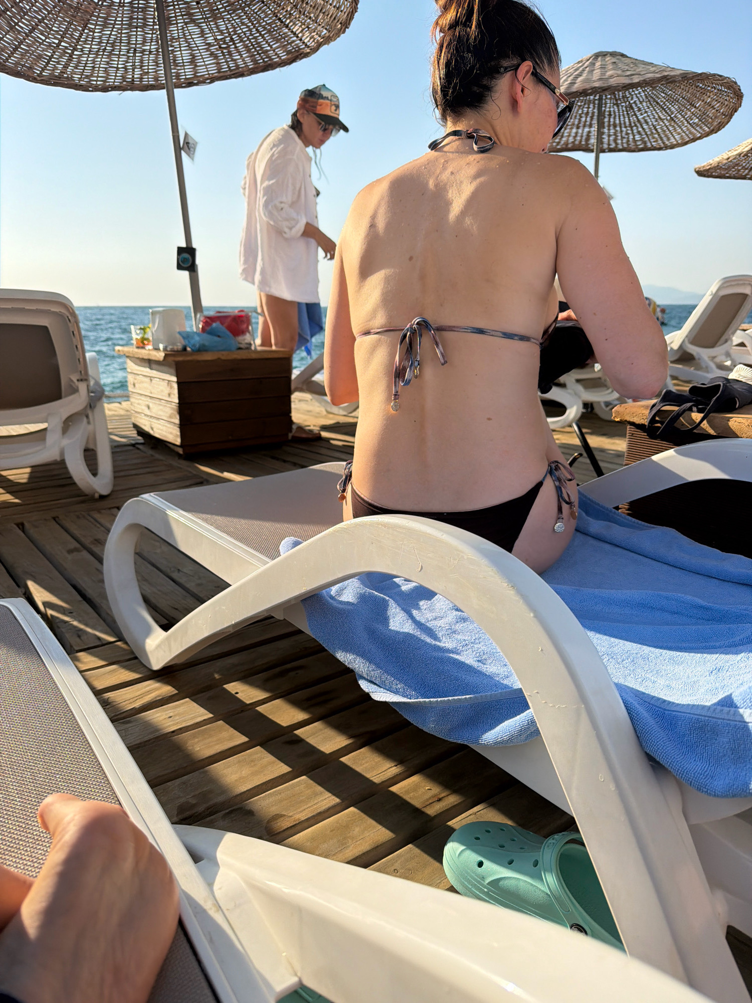 The image depicts a sun-drenched outdoor scene on a wooden deck, likely at a beach or resort in Kuşadası, Turkey. In the foreground, a person is partially visible, with their hand and foot resting on a lounge chair covered with a blue towel. Another lounge chair is in the lower left corner. The main subject of the image is a woman from behind, wearing a bikini and sunglasses, relaxing on a lounge chair under a straw umbrella. Her back and shoulders are tanned, and she has a hair bun. To the left, under another umbrella, another person in a white shirt and a baseball cap is standing near a wooden crate holding some items, possibly drinks. The sea is visible in the background, with a clear blue sky overhead. The sunlight casts distinct shadows on the wooden deck, indicating a bright, sunny day. A pair of light blue Crocs are visible under a lounge chair.