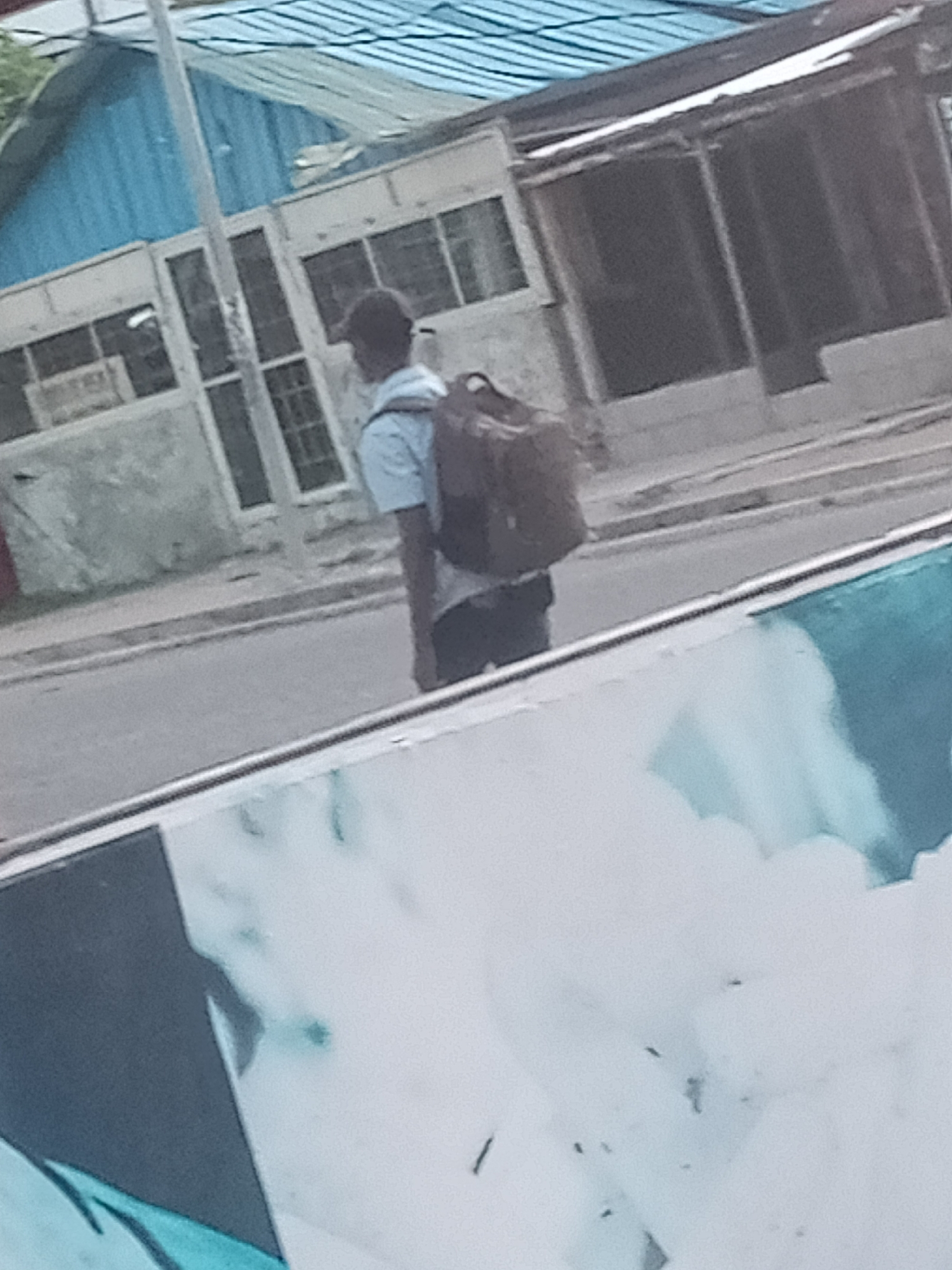 The image shows a person wearing a light blue shirt and dark pants, with a dark backpack, walking on a paved road in Jalingo, Nigeria. The person is facing away from the camera and appears to be walking towards the left of the frame. In the background, there are buildings with corrugated metal roofs and what appear to be storefronts with windows. A utility pole is visible to the left of the person. The foreground of the image is dominated by a partially visible structure with a white surface and some blue abstract shapes. The overall scene depicts a street view in an urban setting.