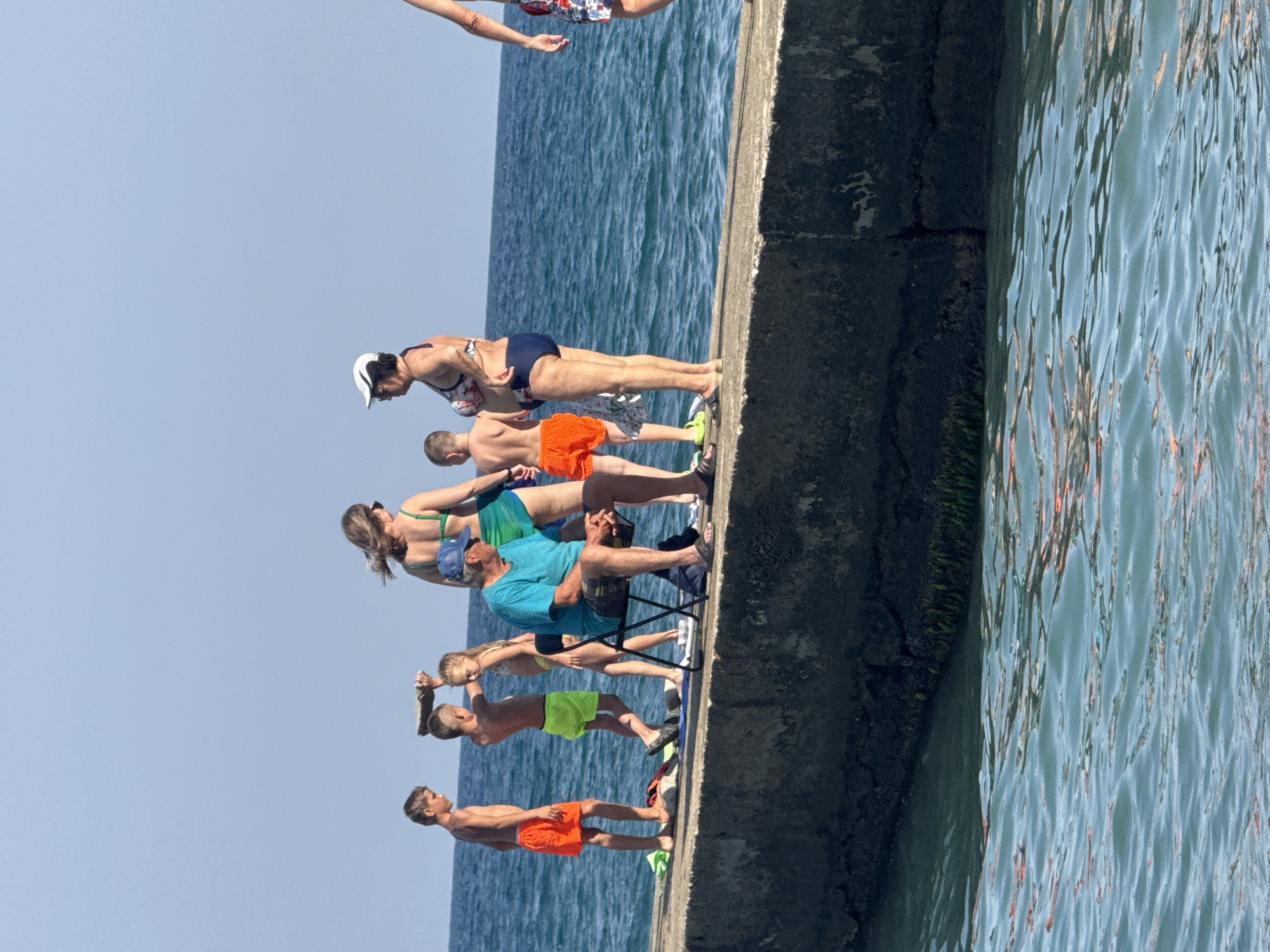 In Partenit, Ukraine, on a sunny day, a group of people are gathered on a concrete pier extending into the Black Sea. The water is a deep blue with visible ripples. The pier is dark gray and appears weathered.
On the pier, several individuals are present. On the left, a boy in orange shorts stands near the edge. Next to him, another boy in neon green shorts holds a flat object above his head, seemingly for balance. A young girl in a yellow bikini stands beside the second boy.
A man in a bright turquoise t-shirt and plaid shorts sits in a black folding chair, facing the sea. To his right, a woman in a green bikini and a woman in a patterned bikini with a white cap are engaged in conversation. Further to the right, partially visible, is a person wearing a patterned swimsuit.
The sky is a clear, light blue, indicating good weather. The overall scene depicts a relaxed atmosphere of people enjoying a day at the seaside.