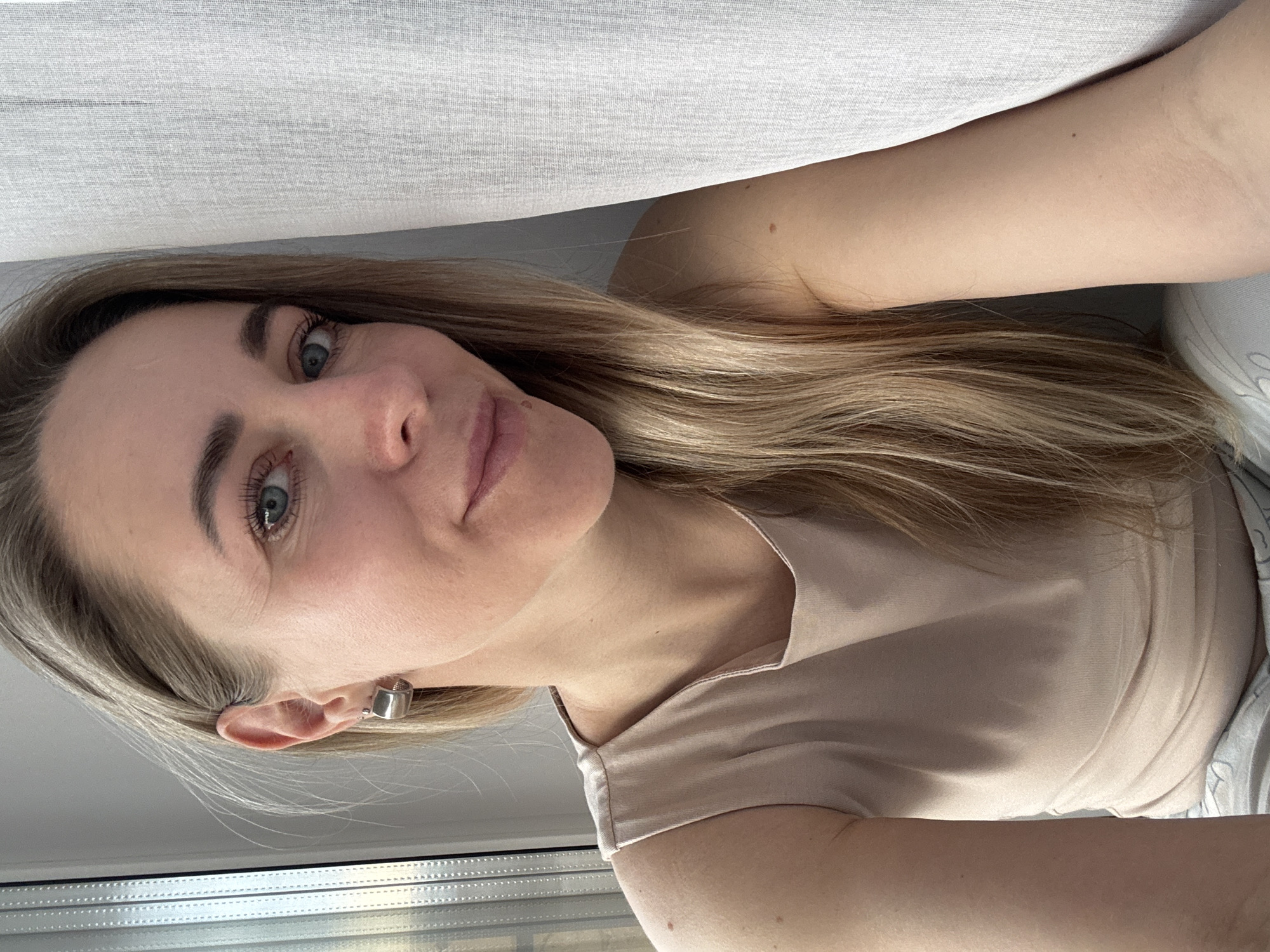 A selfie photograph depicts a woman indoors near a window. The woman has light blonde hair, fair skin, and blue eyes. She is wearing a light beige top and a silver earring on her left ear. Part of a window with a white frame is visible at the bottom, and a white curtain is visible at the top of the frame. The location is assumed to be in Chaikovsky, Russia, based on the prompt instructions, although no specific details in the image confirm this.