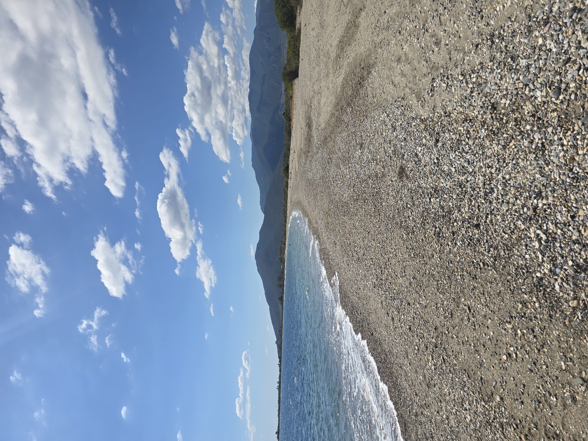 A wide, pebble-strewn beach stretches along the coast of Astros, Greece, under a bright blue sky dotted with white, fluffy clouds. The sea, a deep blue, gently laps at the shore, creating a thin line of white foam. In the distance, rolling green mountains form a scenic backdrop, their peaks softened by the haze. The beach itself is a mix of sand and countless small, smooth pebbles in various shades of grey, white, and brown. A few scattered green bushes and low-lying vegetation can be seen along the edge of the beach, hinting at the natural landscape of the region. The scene is serene and peaceful, evoking a sense of a quiet summer day by the sea.