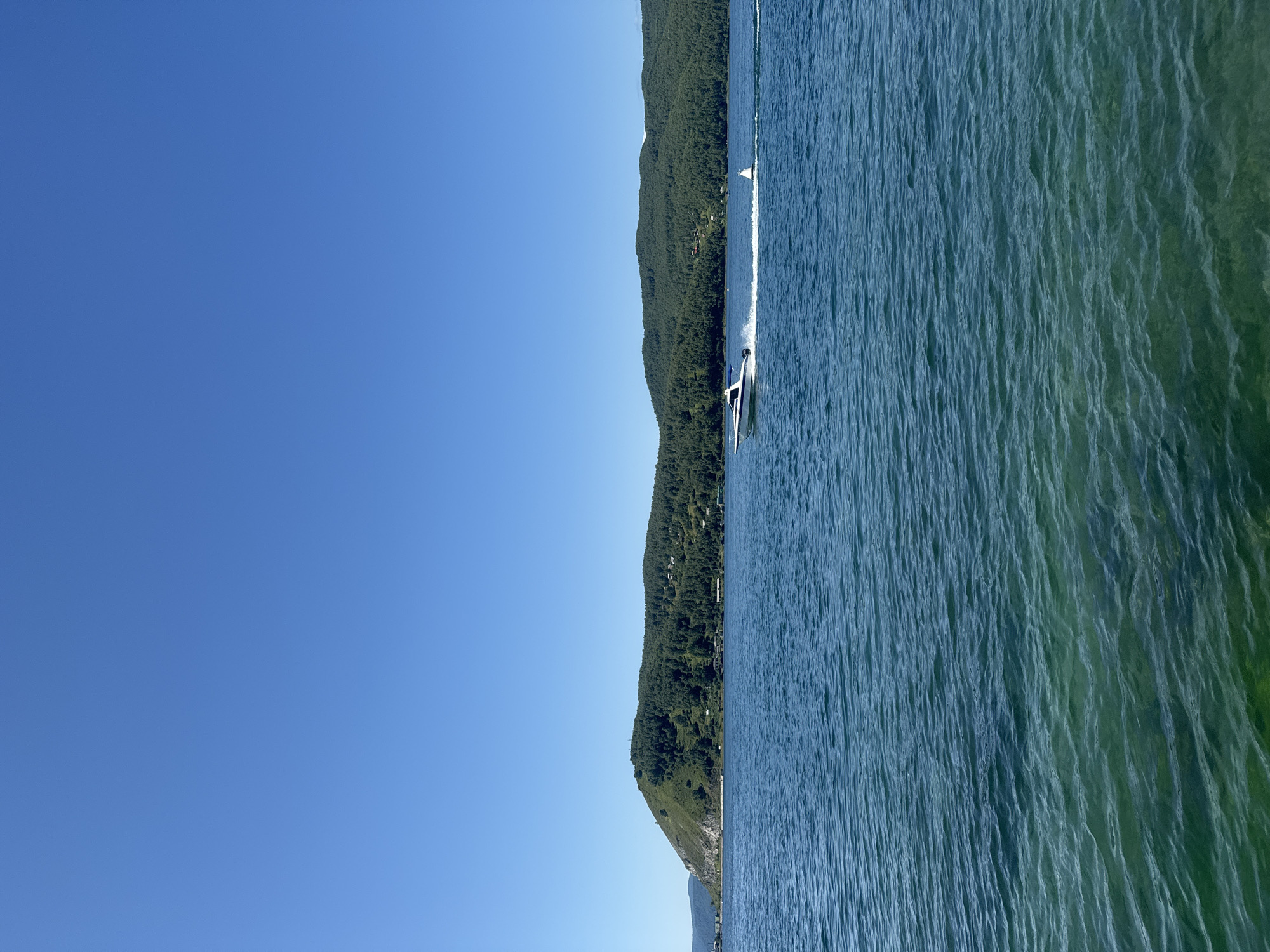 A motorboat speeds across a vast blue lake under a clear blue sky. The boat, white with a blue stripe, leaves a white wake behind it. In the distance, a tree-covered hill rises from the water's edge, with scattered buildings visible among the trees. The lake water is a deep blue in the distance and becomes a translucent green closer to the foreground, revealing the rocky bottom. A white buoy is visible in the water to the right of the boat. The scene appears to be on Lake Baikal, near Listvyanka, Russia, a popular tourist destination known for its clear waters and surrounding natural beauty.