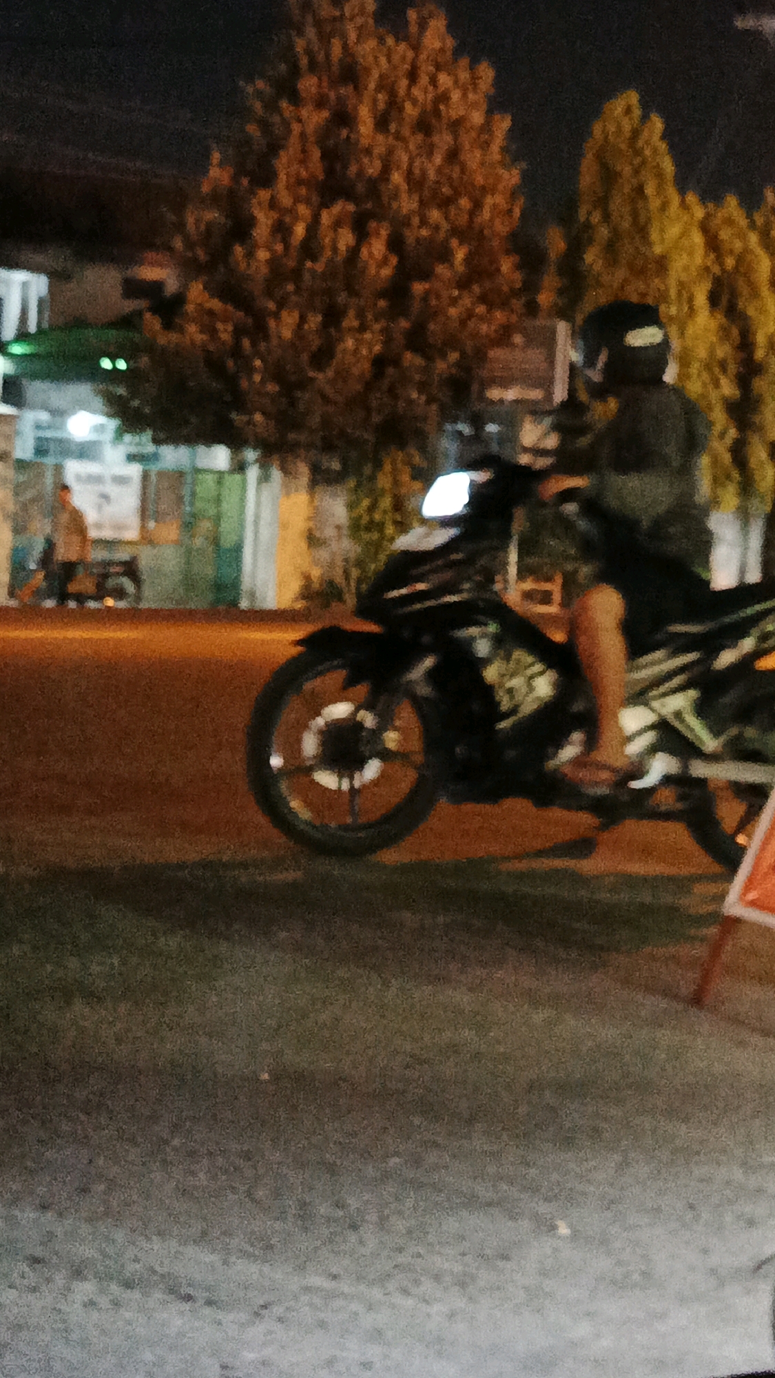 The image captures a nighttime scene on a street in Surakarta, Indonesia, featuring a person riding a motorcycle. The rider, wearing a dark helmet and a dark long-sleeved shirt, is positioned on a black motorcycle with white accents and a bright headlight illuminated. The motorcycle's front wheel, with a silver rim and visible spokes, is prominent in the foreground. Behind the motorcycle, a street is visible with an orange glow from streetlights or the road surface, and a strip of dark grass or ground in the foreground. In the background, there are several trees with orange and yellow foliage, and the outlines of buildings, one of which appears to have green accents and glowing lights. Another person is standing near a parked motorcycle in the middle distance, to the left of the frame. The overall impression is of a casual, urban nighttime environment.