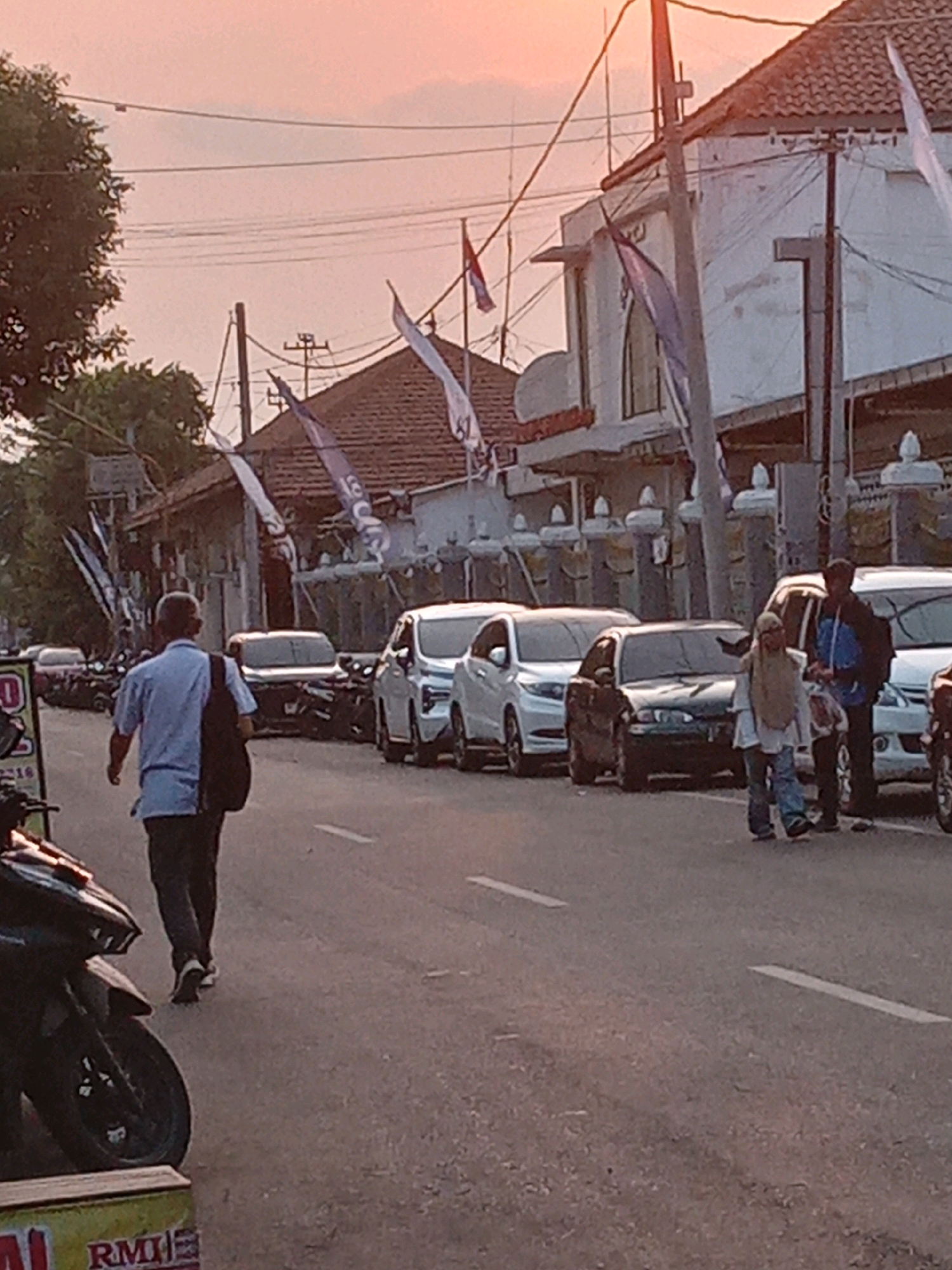 The image depicts a street scene in Yogyakarta, Indonesia, during what appears to be sunset. The sky is a soft orange and pink, indicating the end of the day. A row of parked cars lines the street, with a few people walking and standing nearby.
In the foreground on the left, a dark-colored motorcycle is partially visible. To the right of the motorcycle, a man in a light blue shirt and dark pants is walking away from the camera, carrying a black backpack.
Further down the street, a line of cars is visible, including a white sedan, a dark-colored sedan, and a white SUV. Two people, a woman wearing a hijab and a man in a blue jacket, are standing near the white SUV.
The buildings along the street have traditional Indonesian architecture with tiled roofs. Several flags are displayed on poles in front of the buildings. The overall impression is a typical street scene in a residential or commercial area of Yogyakarta, with the warm glow of sunset creating a tranquil atmosphere.