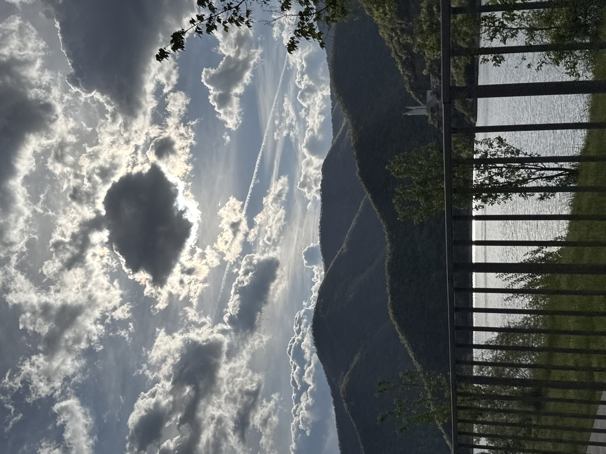 The image shows a landscape with a body of water viewed through a metal railing. To the left are mountains partially obscured by a cloudy sky with contrails. A dark cloud obscures the sun. The location is Mihalkovo, Bulgaria, inferred from metadata.