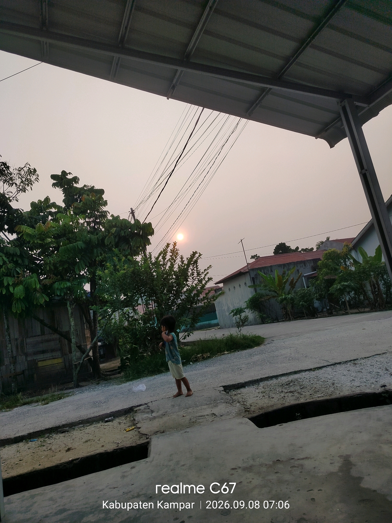 A young child stands on a paved road in what appears to be a residential area at dusk. The child is wearing a light-colored t-shirt and shorts. In the background, there are houses with red roofs and lush green vegetation. Power lines are visible crisscrossing the sky, and the sun is setting, casting a warm glow. The foreground shows a drainage ditch. The image is dated 2026.09.08 at 07:06, and the location is identified as Kabupaten Kampar, Indonesia.