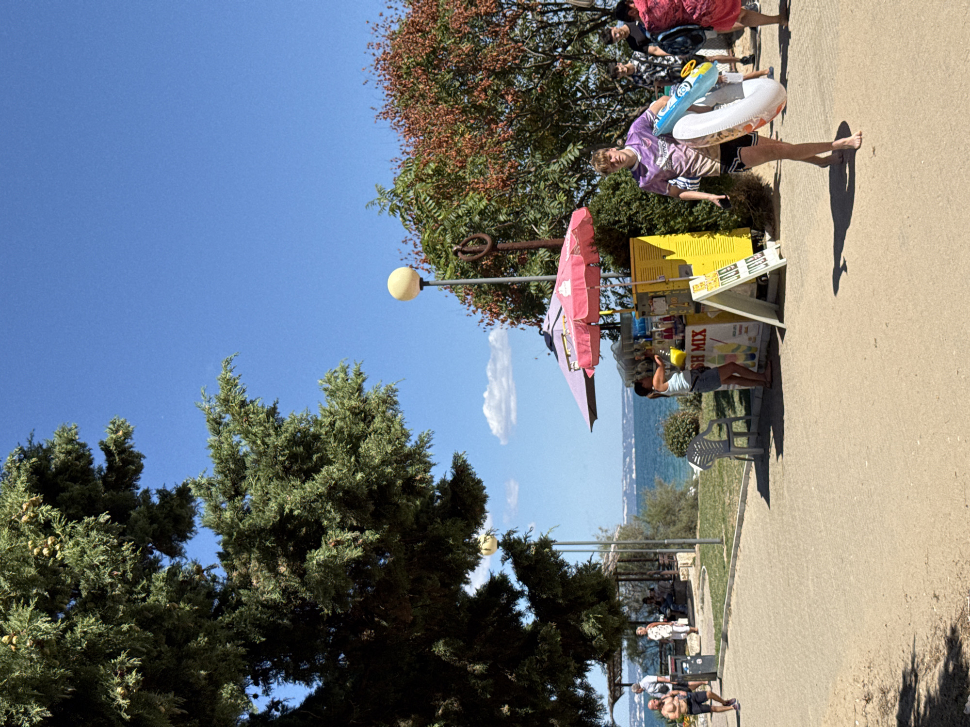 The image depicts a sunny day at a seaside location in Varna, Bulgaria. A paved walkway runs in the foreground, with sand visible on the left. To the right, a young man wearing a purple t-shirt and black shorts walks barefoot, carrying a blue and white inflatable ring. Behind him, other people are visible, including individuals near a yellow structure with a pink umbrella, likely a kiosk selling drinks or snacks. A small white chair is placed near the kiosk. In the background, a body of water, presumably the Black Sea, stretches to the horizon with a coastline visible in the distance. Large trees with dense foliage, one on the left with hanging cones, and another with reddish-brown leaves on the right, frame the scene. The sky is clear and blue with a few scattered clouds. The location appears to be a popular recreational area along the Varna coast.