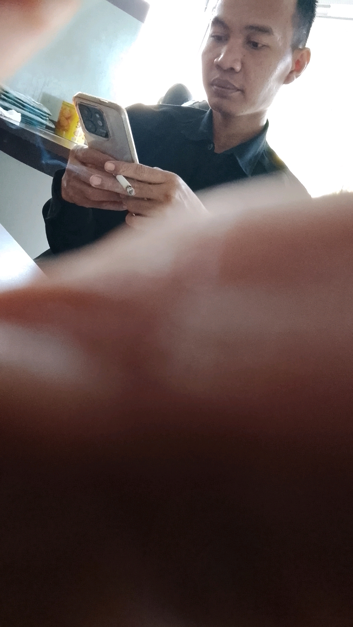 An Indonesian man in a black collared shirt is holding a smartphone in his left hand and a lit cigarette in his right hand. The cigarette is actively burning and emitting smoke. The man is looking down at his phone. In the background, there are stacks of papers and a brightly lit window. The foreground is blurred, obscuring a significant portion of the image. The scene is likely indoors, possibly an office or a home in Slawi, Indonesia.
