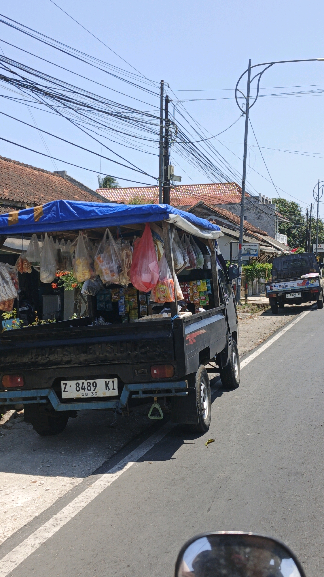 A black pickup truck, modified into a mobile store, is parked on the side of a paved road in Cikembulan, Indonesia. The truck's bed is covered by a blue tarp, beneath which various items are displayed and hung in plastic bags. These include snacks, drinks, and other retail goods. The truck has a license plate that reads "Z 8489 KI". In the background, there are buildings with red tile roofs, utility poles with a dense network of electrical wires, and a clear blue sky. Another pickup truck is visible further down the road. The scene appears to be a sunny day.