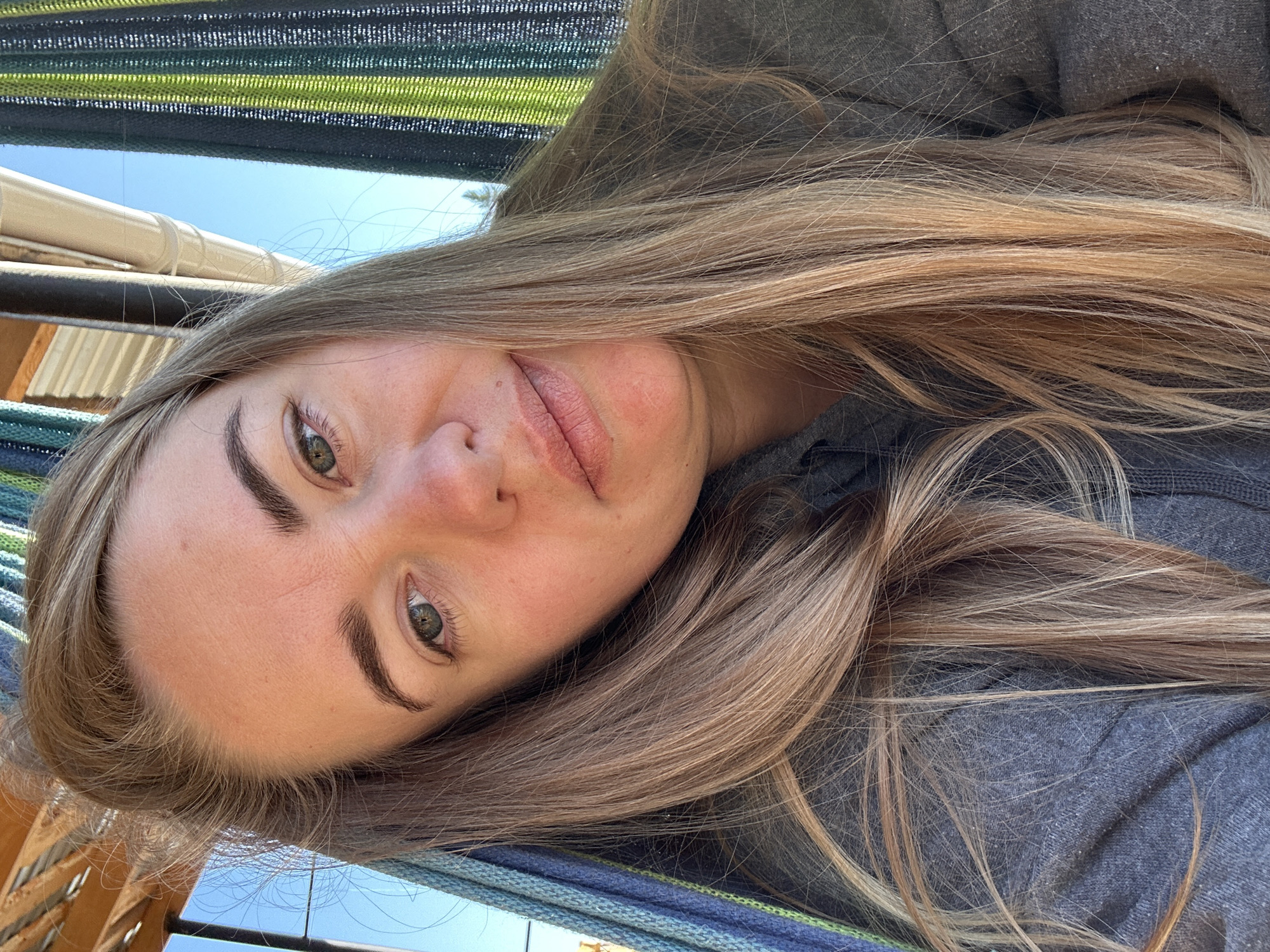 A woman with long, light brown hair is lying in a hammock. Her face is turned towards the camera. She has fair skin, green eyes, and defined eyebrows. She is wearing a gray top. The hammock is made of green and blue striped material. The sky is visible in the background. The location is not determinable from the image.