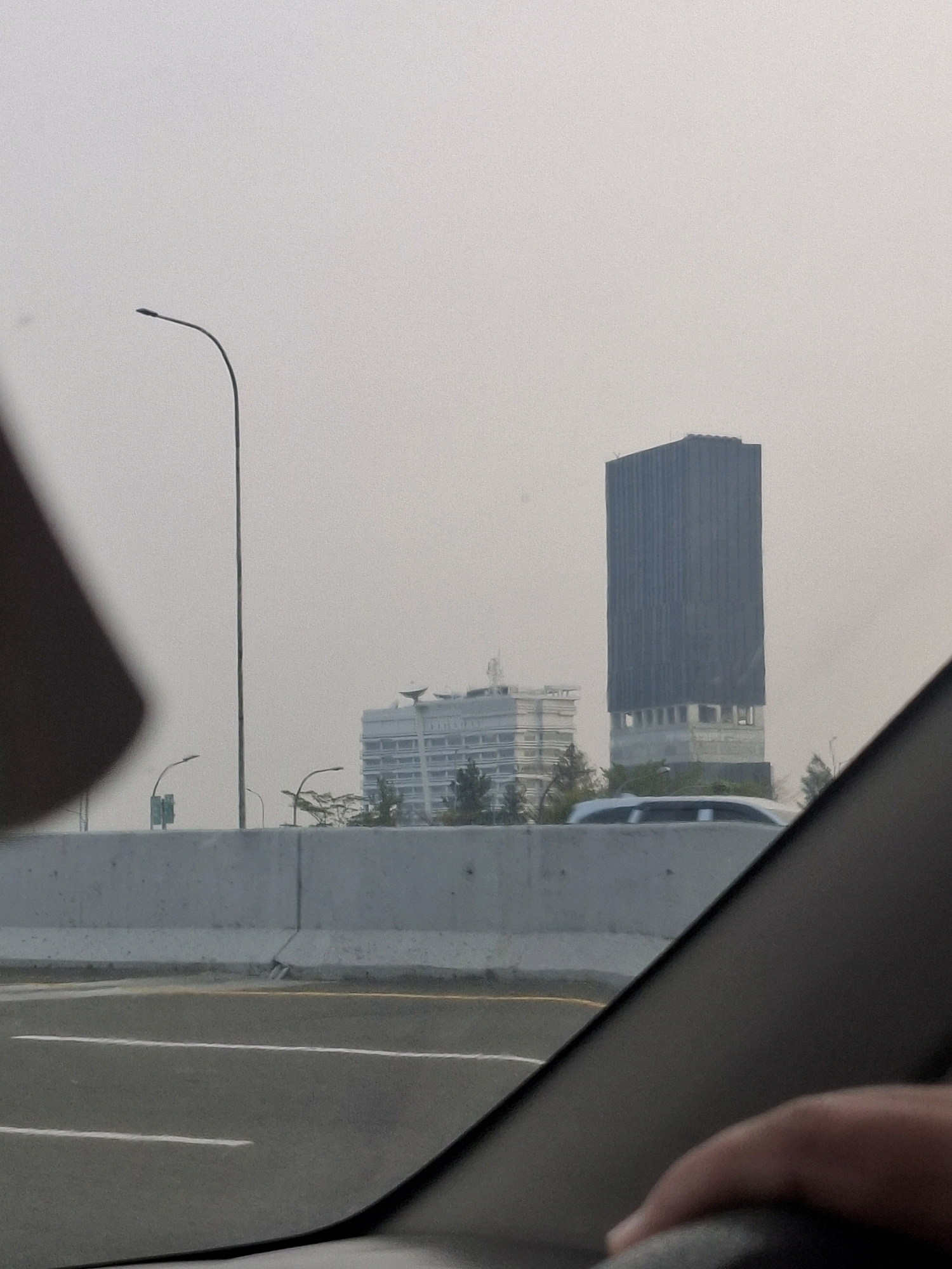 The image shows a view from inside a car on a highway in Tangerang, Indonesia. In the background, there are two tall buildings and a few smaller structures. The sky is hazy, suggesting an overcast day or possibly air pollution. A concrete barrier separates the highway from the area where the buildings are located. There are streetlights visible along the highway. A hand is visible on the steering wheel in the foreground, indicating the perspective of the driver or passenger. The overall scene is urban, with a focus on infrastructure and architecture.