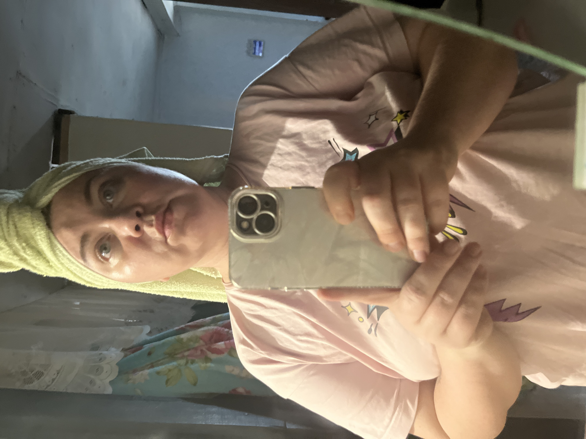 A woman with a light green towel wrapped around her head takes a selfie in a mirror. She is wearing a pink t-shirt with a graphic print of stars and lightning bolts. She is holding a silver smartphone with a multiple camera system in her right hand. The background appears to be a bathroom, with a floral curtain visible on the left and a white ceiling with visible cracks. The lighting is dim, suggesting it is either early morning or evening. The image was taken in a residential bathroom in the city of Fokino, Russia.