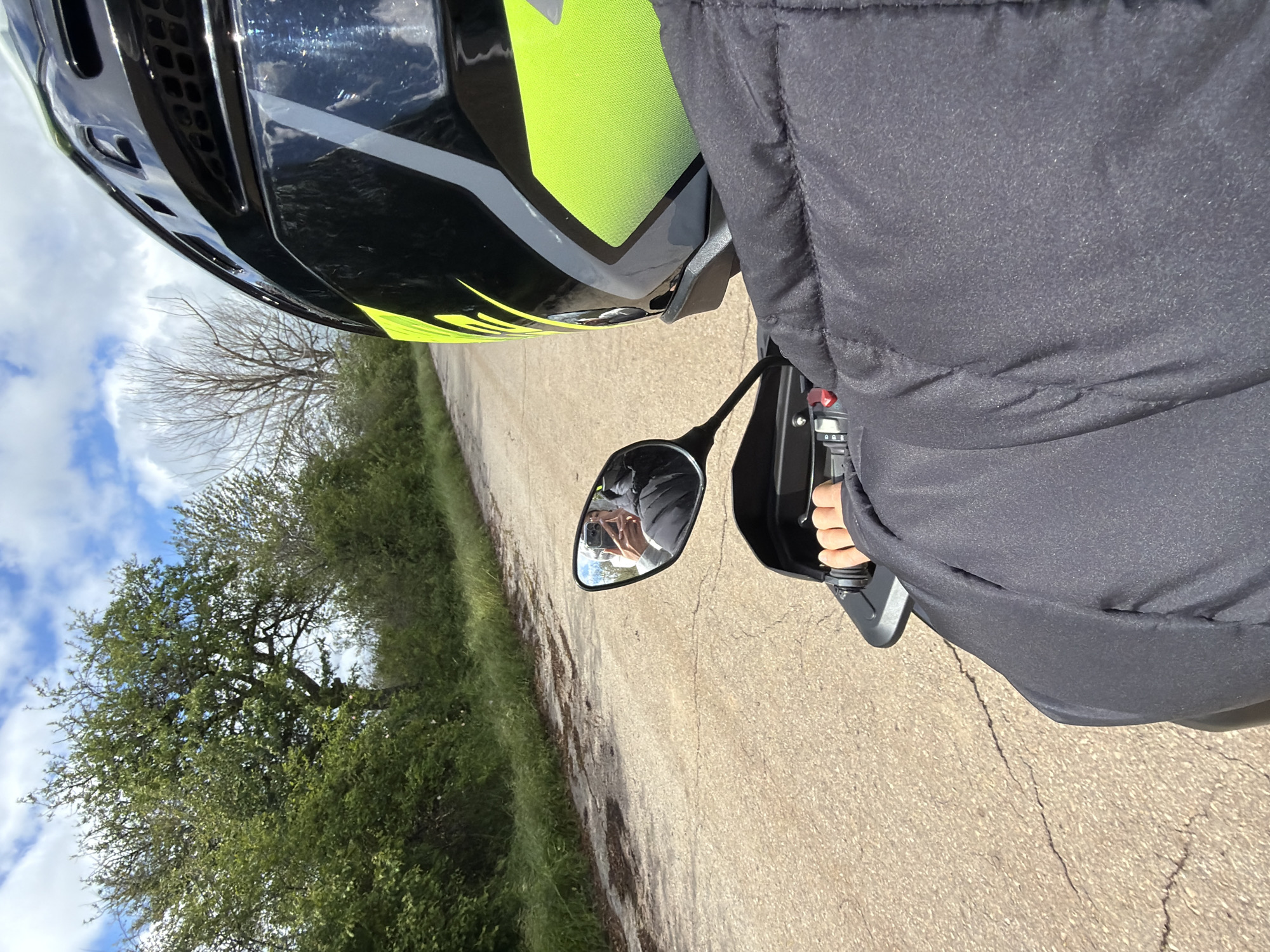 The image shows a motorcyclist wearing a black and neon green helmet and a black jacket riding on a paved road in what appears to be a rural area. The rider's hand is visible on the motorcycle's handlebar, and the rearview mirror reflects the rider taking a picture with a smartphone. A tree line and cloudy sky are visible in the background. The location is likely near Мичиковци, Bulgaria, as specified.