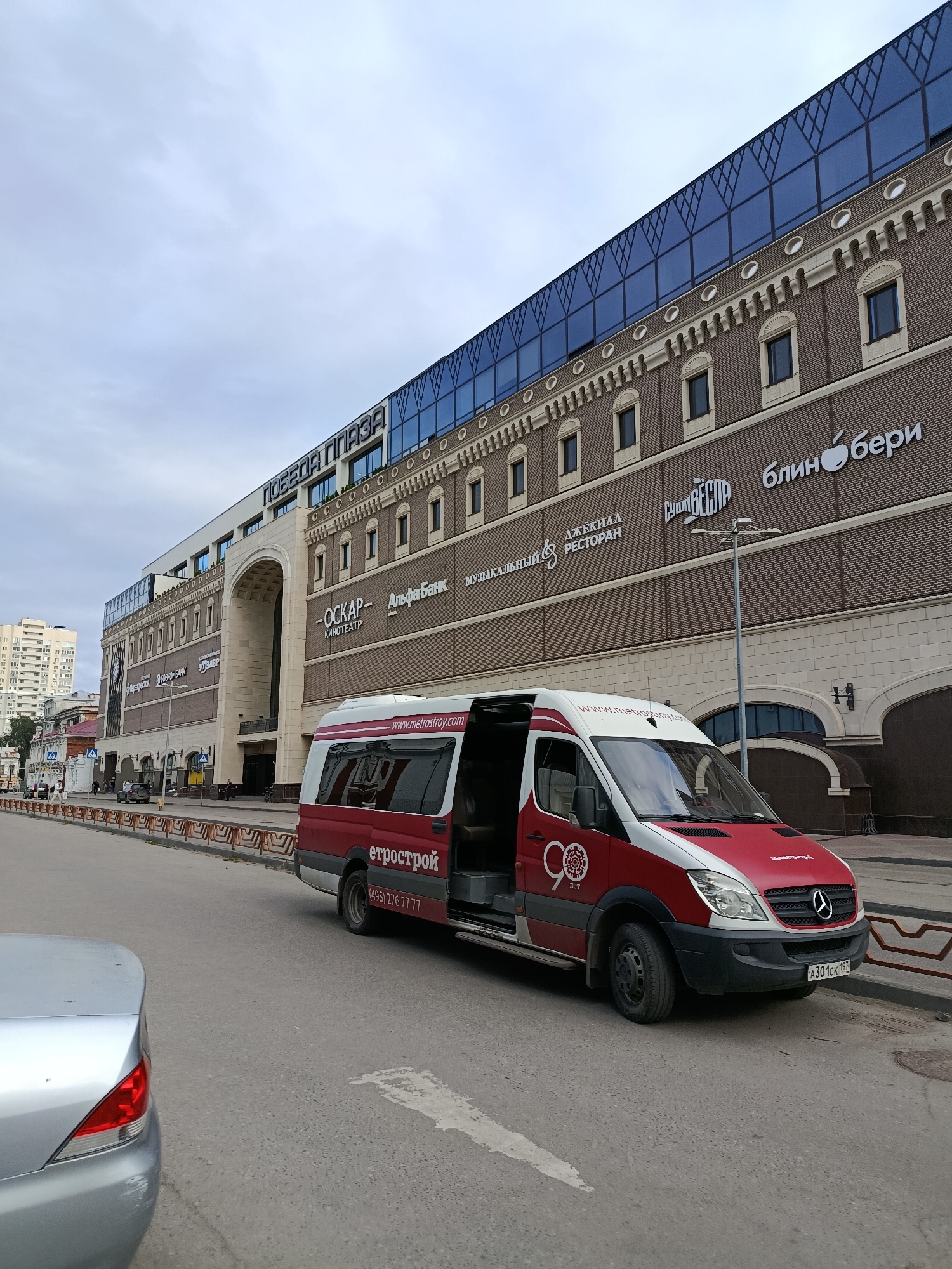 In Saratov, Russia, a red and white Mercedes-Benz Sprinter van with an open side door is parked on a street in front of a large brick building with a modern glass facade. The building has various signs, including "ПОБЕДА ПЛАЗА" at the top, and other advertisements for shops and businesses like "ОСКАР", "АльфаБанк", "Музыкальный", "РЕСТОРАН", "СУШИ ВЕБЕР", and "БлинБери". The van displays a logo with "90 лет" and the website "www.metrostroy.com". The license plate of the van is "A301CK 19". A silver car is partially visible in the foreground on the left. The sky is overcast.