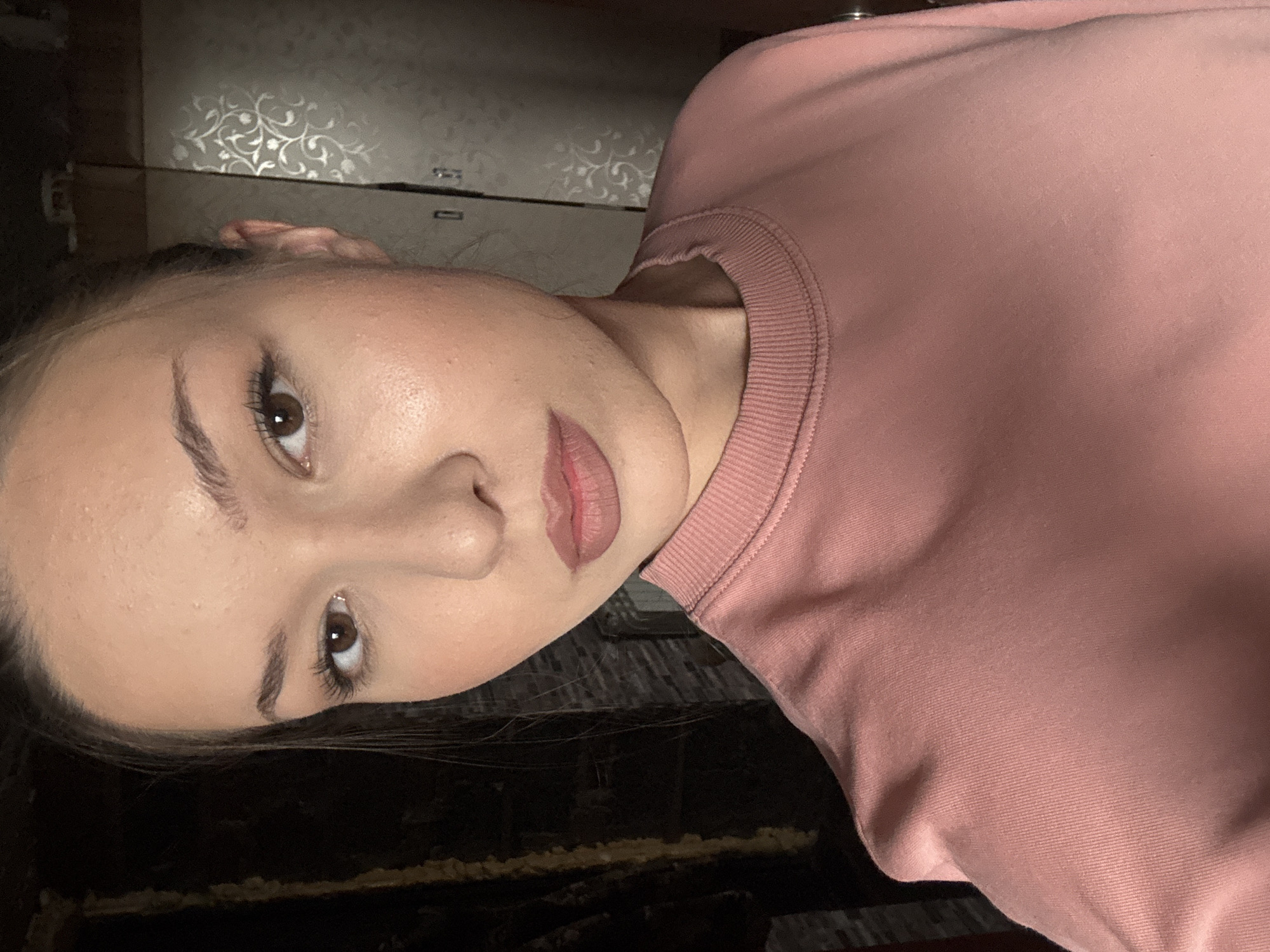 A young woman is captured in a close-up, front-facing selfie, with her face taking up the majority of the frame. She has light skin with a subtle rosy blush on her cheeks and a matte, pinkish lipstick. Her eyes are brown and appear to be wearing mascara and eyeliner. Her dark hair is pulled back. She is wearing a pink crewneck sweatshirt. The background is dimly lit and appears to be an indoor setting, with a patterned wallpaper visible on the right side and a dark, textured wall on the left. The image does not provide any visual information to indicate it is located in Vishnevka, Russia.