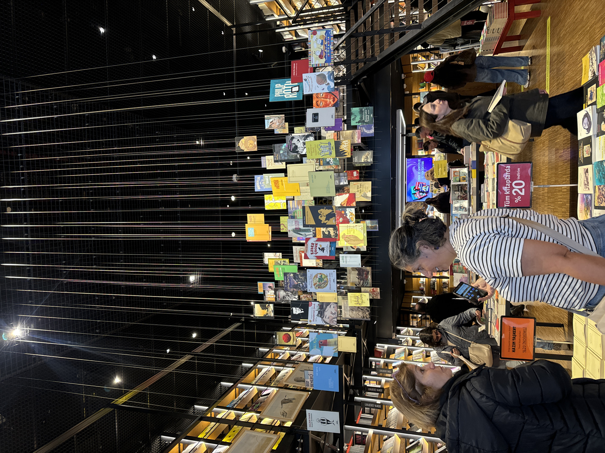 Here is a detailed description of the image:
The image captures the interior of a modern bookstore, likely located in the city of Unknown. The foreground features two women standing close to each other, seemingly engaged in a conversation while browsing books. One of the women is wearing a black jacket. The second woman is wearing a striped top. A sign displays a "20% discount" on all books.
The store has modern interior design. Books are arranged on shelves. Above, a multitude of book covers is suspended from the ceiling by thin lines, creating a visually striking display. The lighting is bright, illuminating the books and the overall space. Other patrons are visible in the background, browsing books on the shelves. The store appears to be busy.