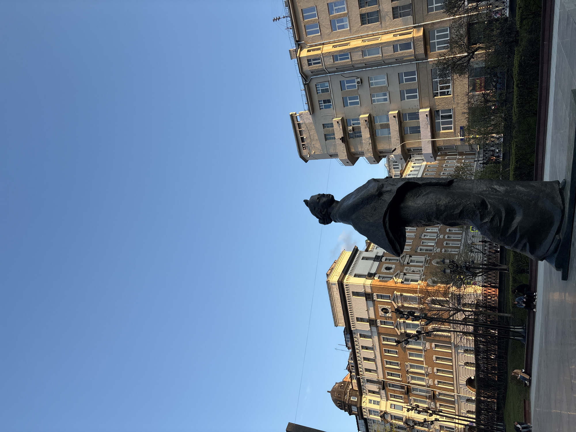 The image shows a bronze statue of a woman, viewed from below and tilted slightly. The statue is positioned in front of buildings with multiple stories and windows. The sky is blue and clear. In the background, there is a fence and some trees. There are people sitting on benches, indicating the statue is in a public area or park. The architecture of the buildings suggests an urban setting likely in Moscow, Russia.