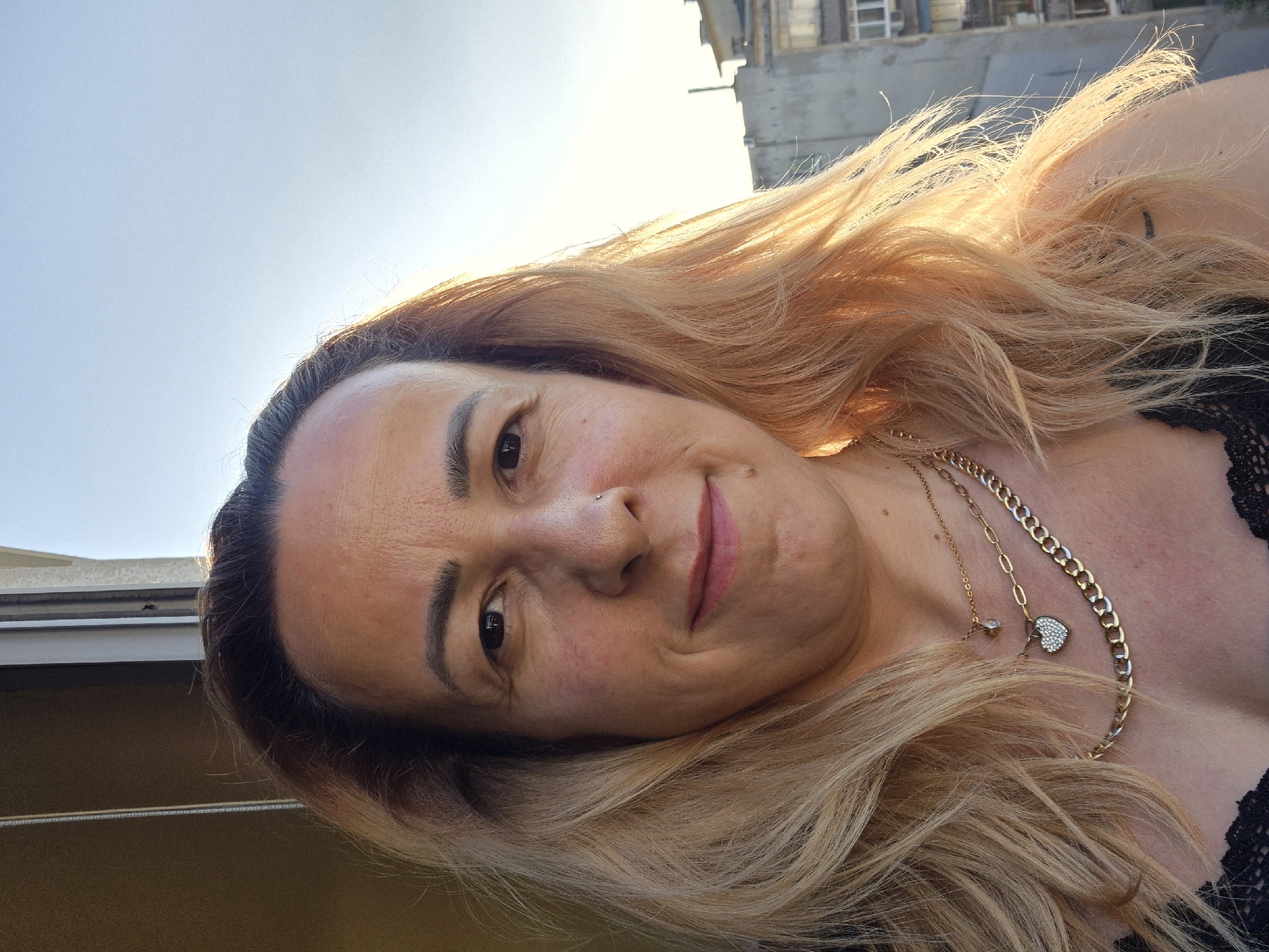 A medium shot portrait of a light-skinned woman with blonde hair is captured outdoors during the day. She is facing forward, with her head slightly tilted. The woman has a small nose ring and is wearing two gold necklaces, one of which has a heart-shaped pendant. Her hair is long and appears to be slightly windswept. In the background, there is a building and a clear sky. The lighting suggests it is either sunrise or sunset, casting a warm glow on her face and hair.